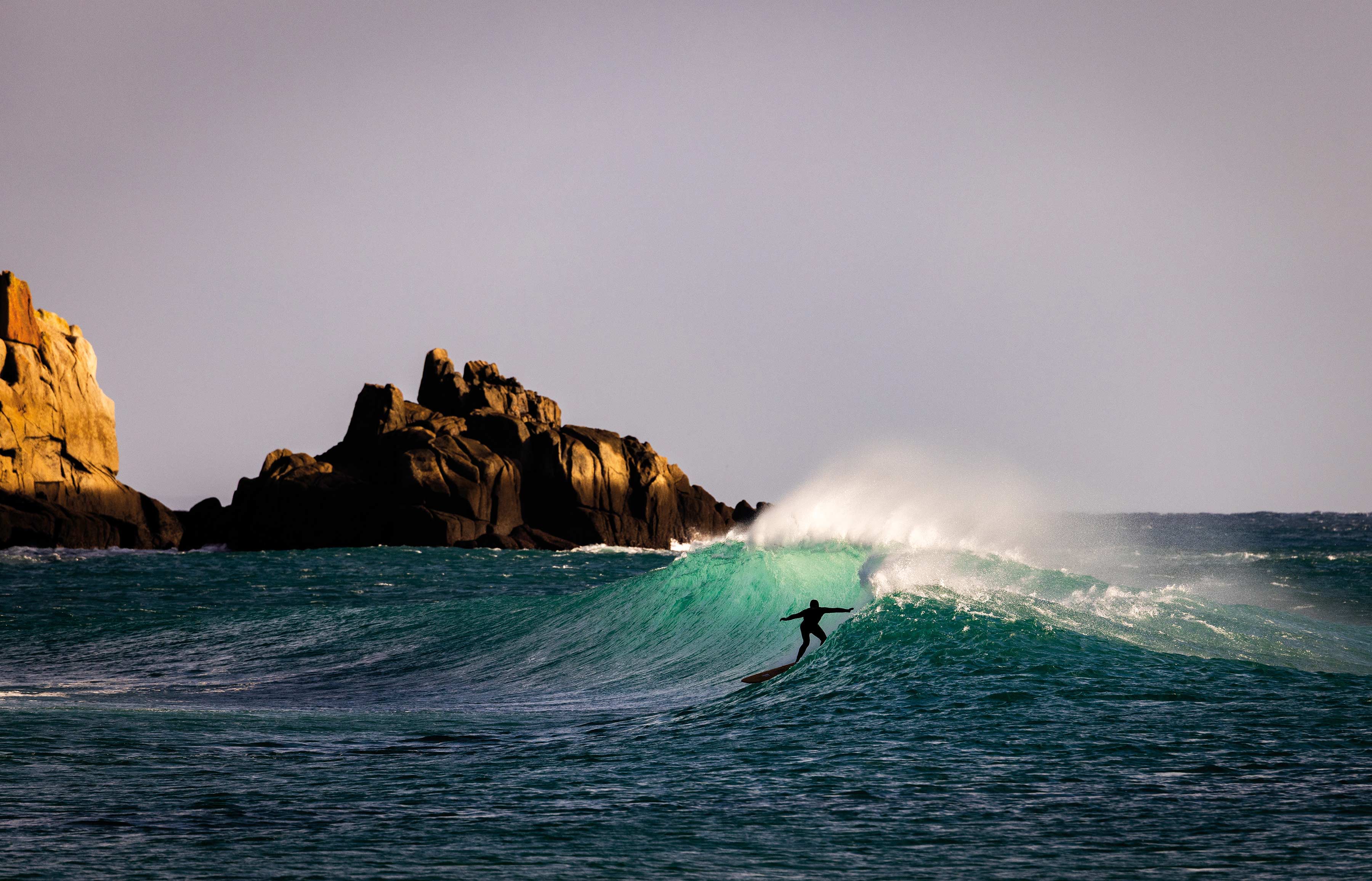 Surfer riding a wave near rocky coastline under a cloudy sky.