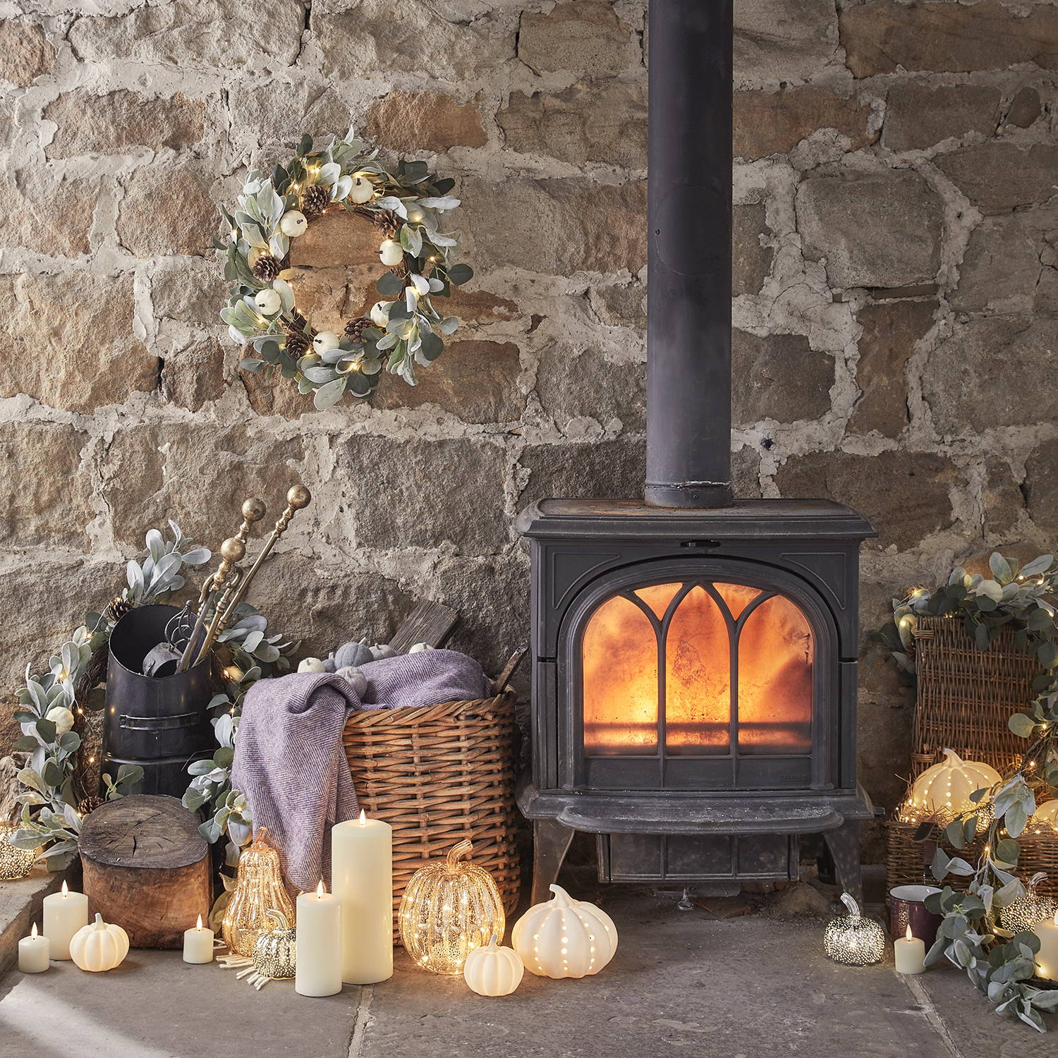 Ein brennender Kaminofen mit danebenstehenden Glaskürbissen, LED Kerzen und verteilten Herbstkränzen und -girlanden