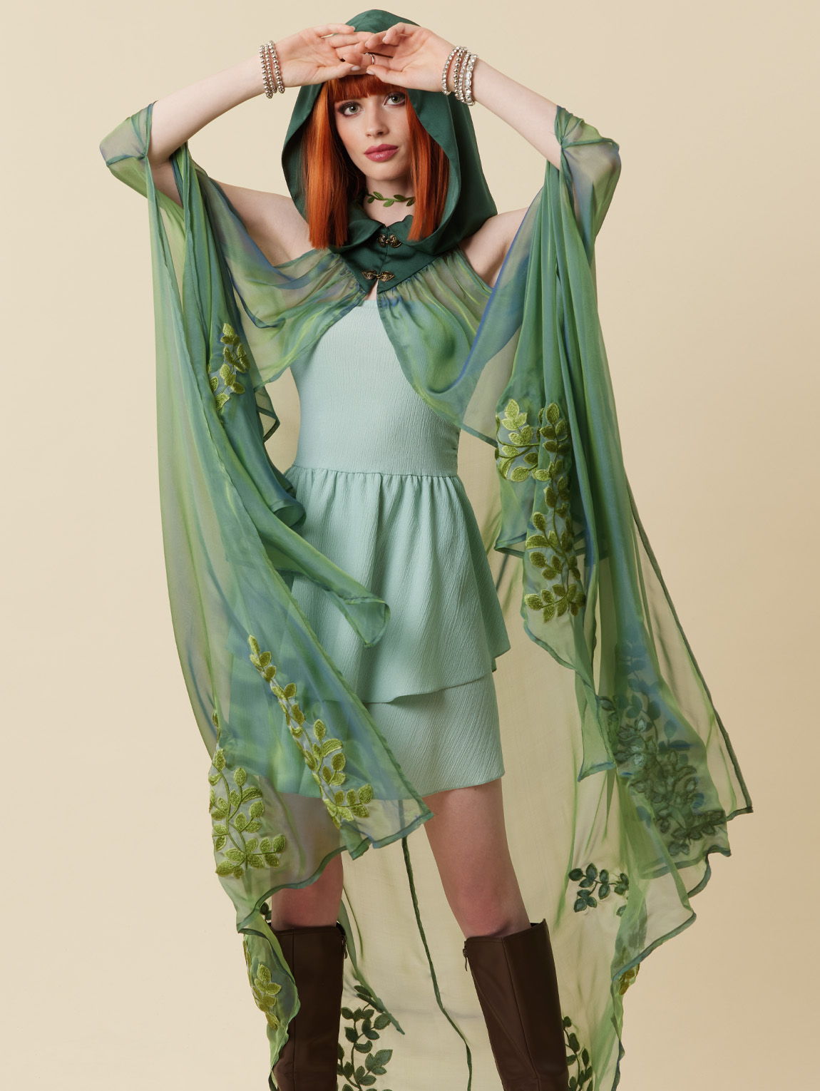Model wearing a green outfit with a flowing cape against a beige background.