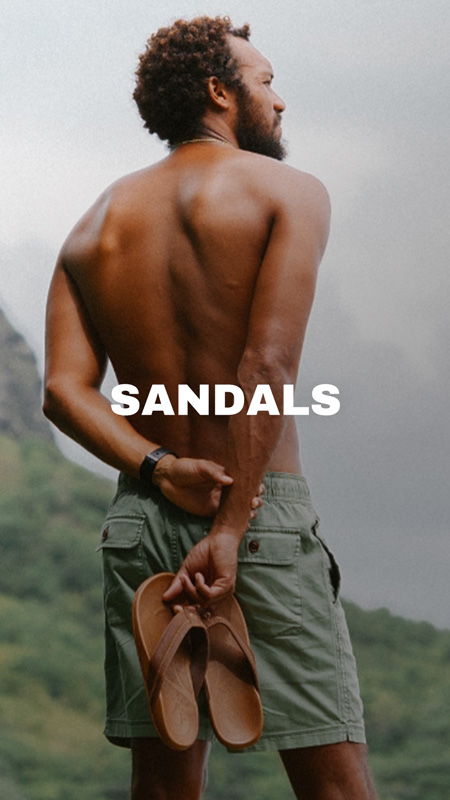 Man holding sandals, standing outdoors with a scenic background.