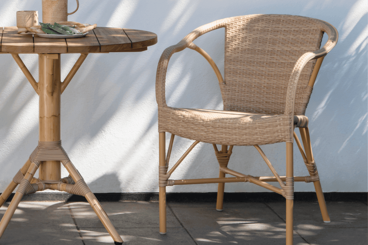 Woven outdoor wicker armchair beside a round teak patio table in dappled shade.