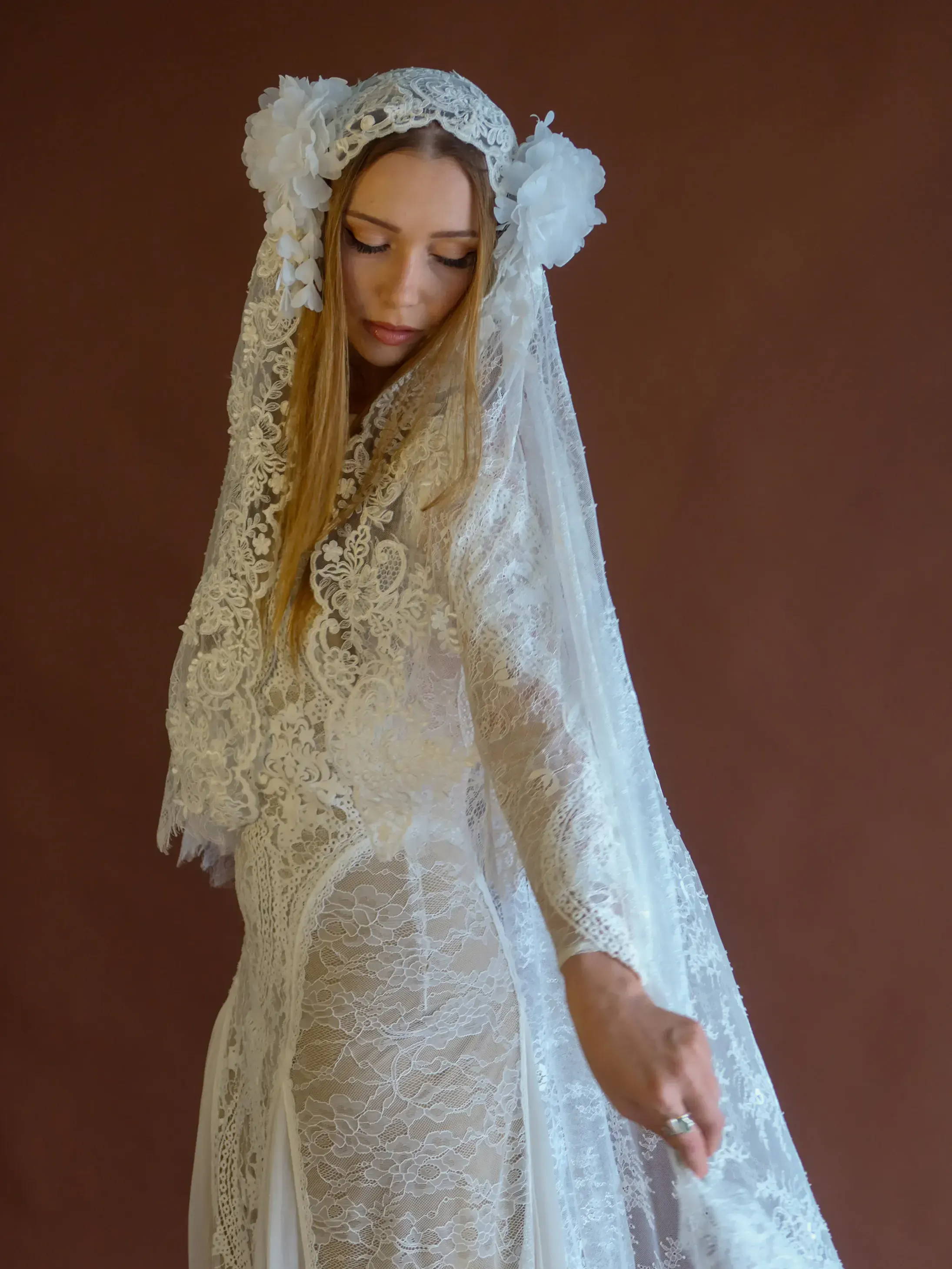 Woman in a white lace dress and veil against a brown background.