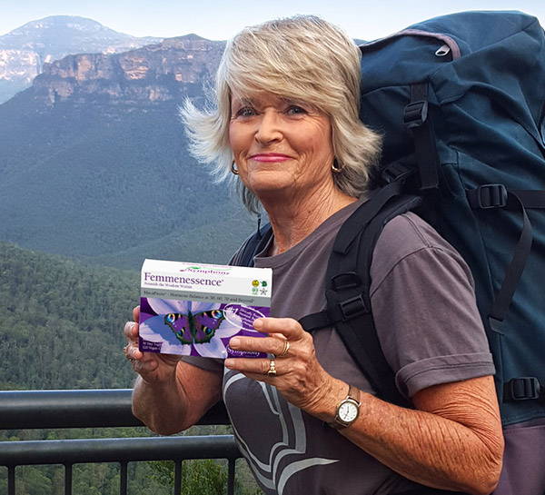 Blond older woman outside with mountains in the background, and wearing a gray t-shirt and carrying a navy backpack while holding Femmenessence MacaPause