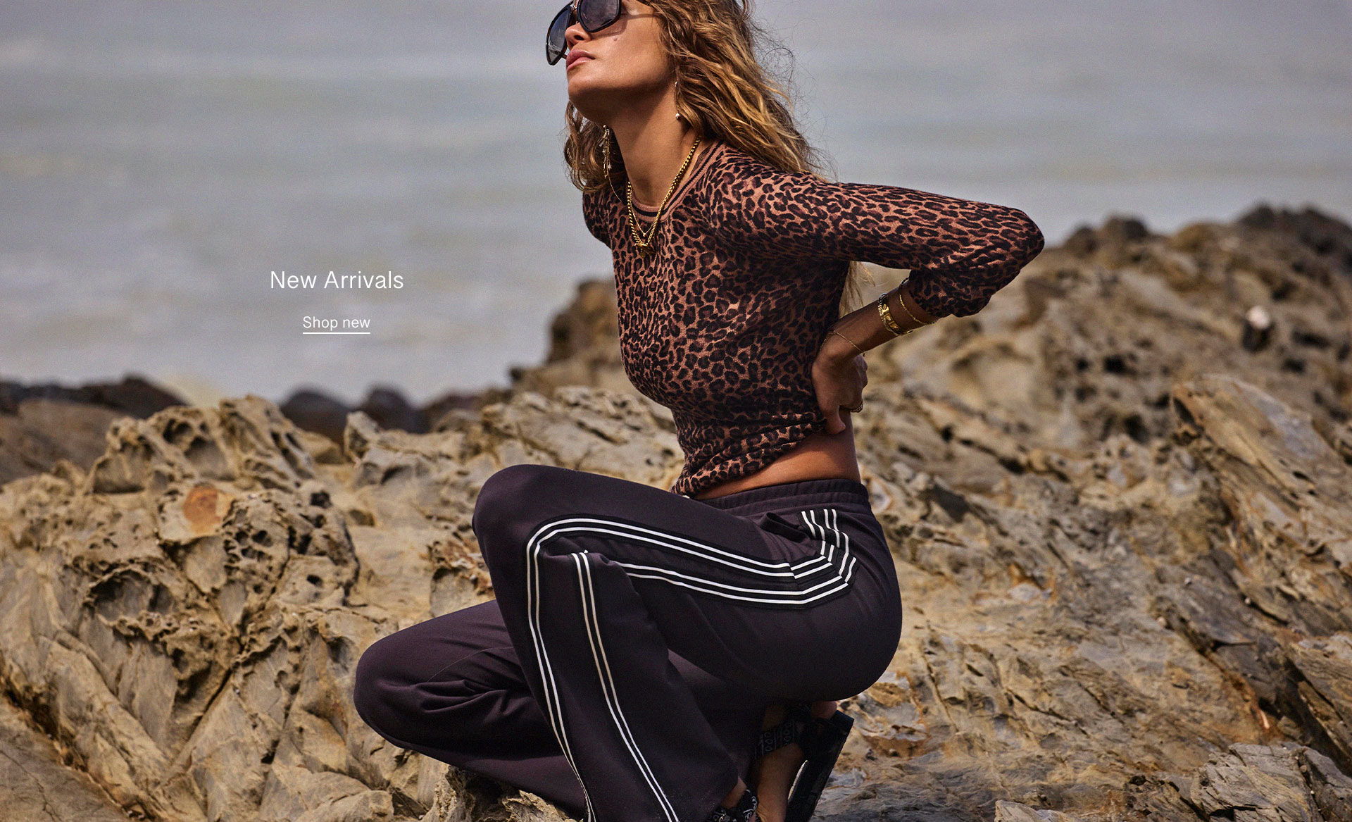 A person in stylish clothing poses on rocky terrain by the sea.