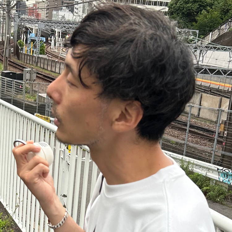 A man with short hair stands outdoors, holding an object and smoking.