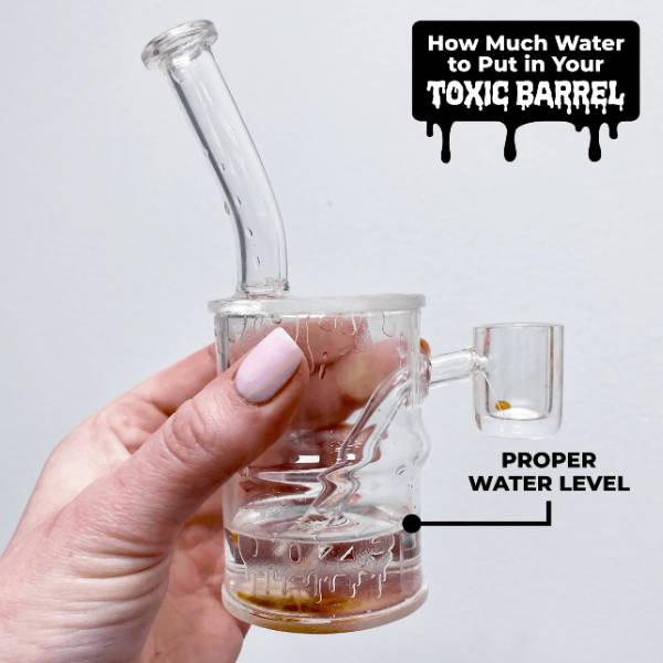 A white girl with light pink nails is holding an Ooze Toxic Barrel quartz mini dab rig that is filled to the proper water level. There is a black arrow pointing to the water level.