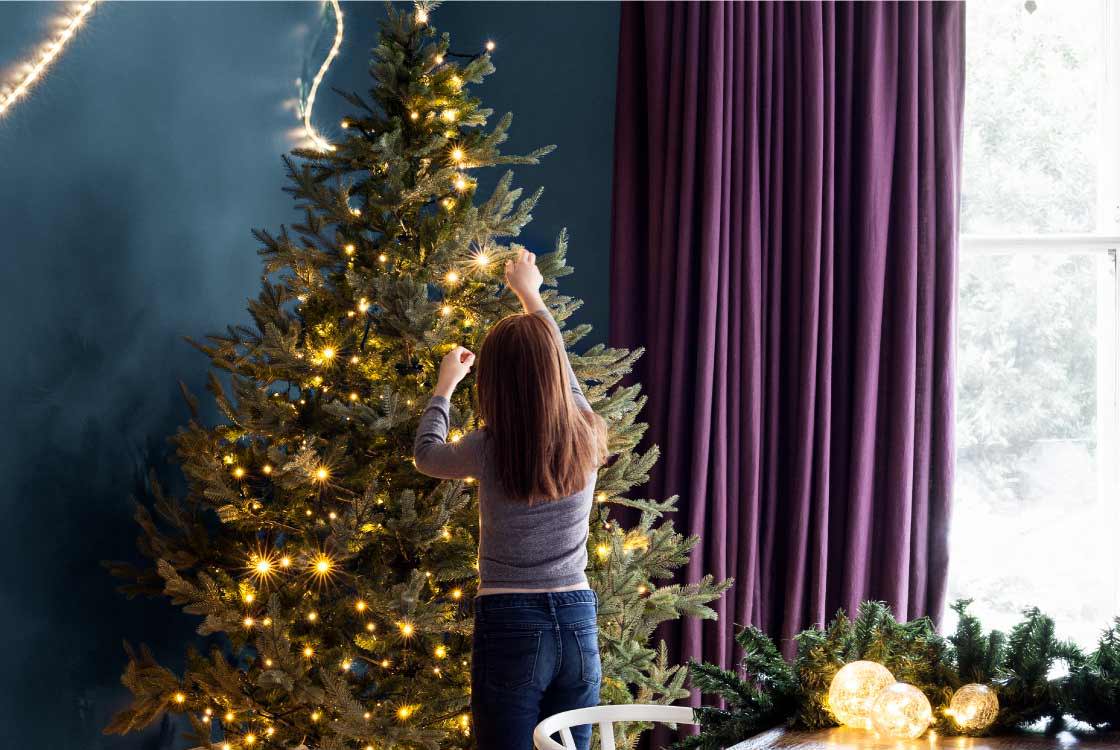 Christmas tree being decorated with a blue wall and burgundy curtains.