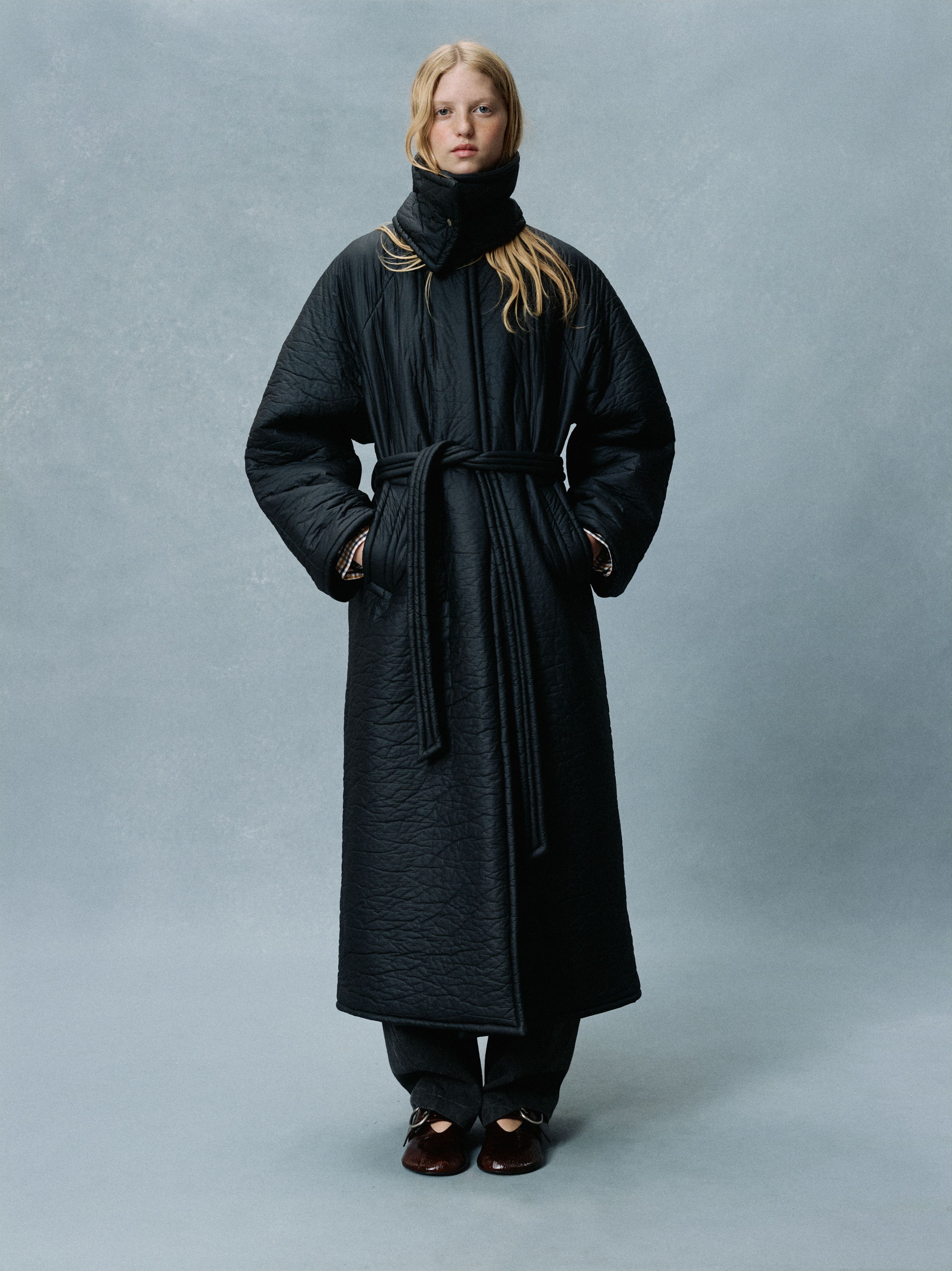 Woman wearing a long black coat, standing against a light blue backdrop.