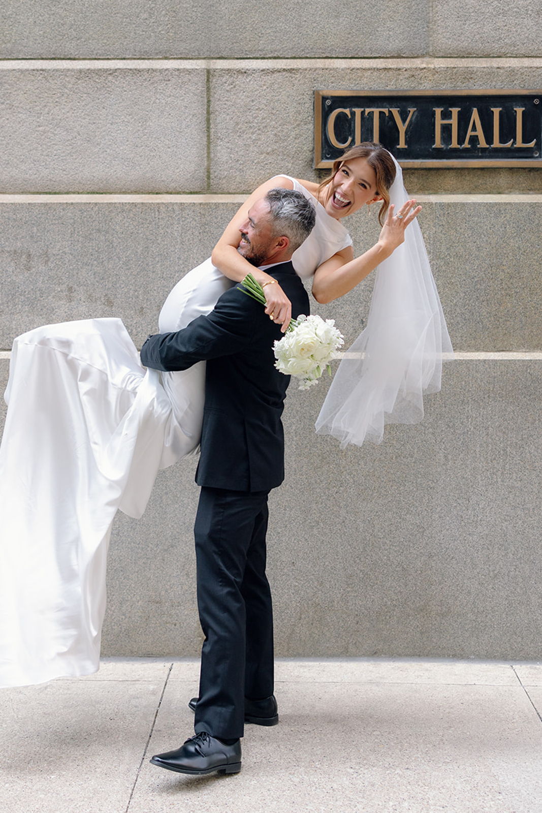 Bride and groom celebrating outside City Hall, with the bride being lifted.