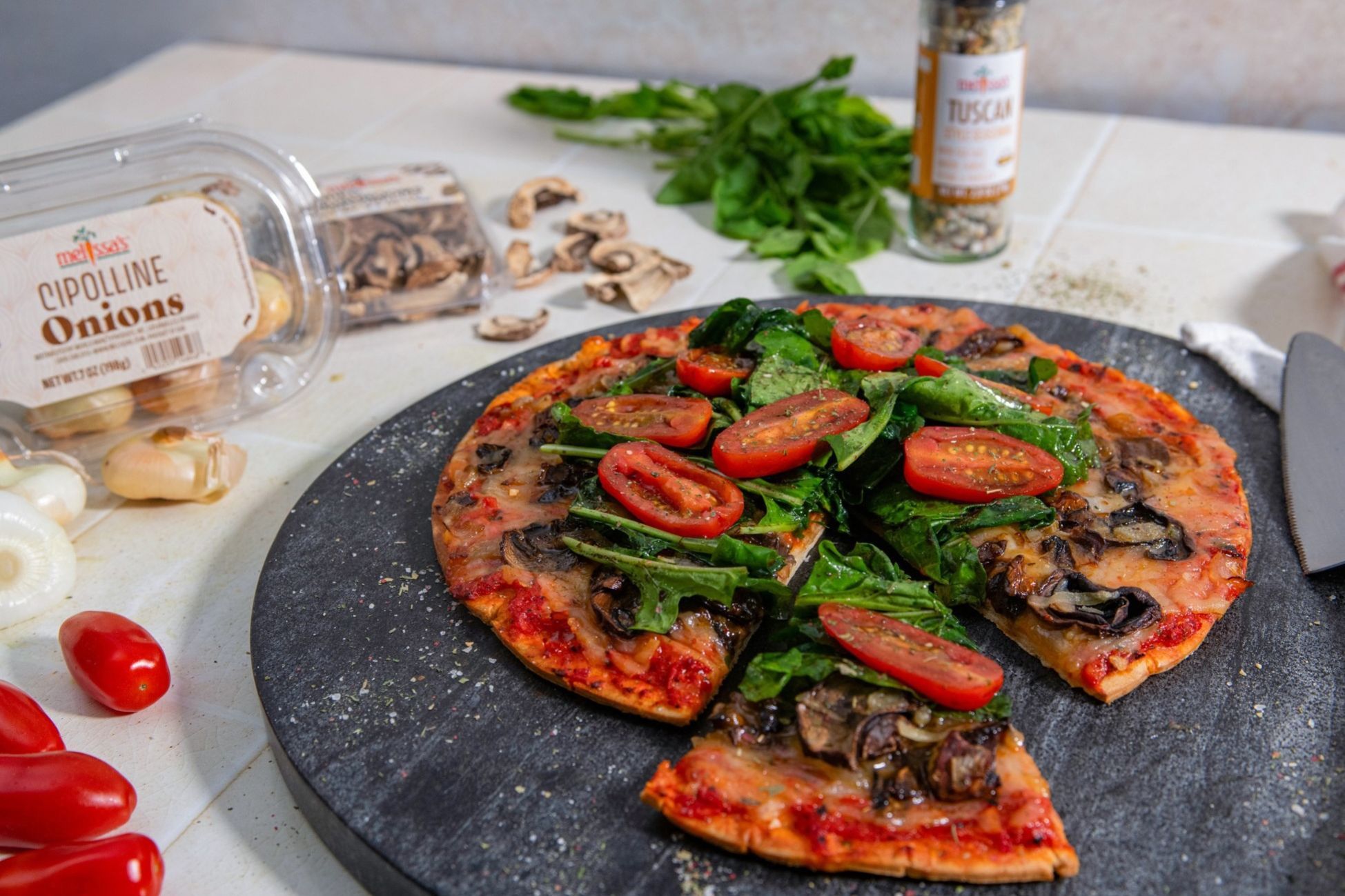 A pizza topped with spinach, tomatoes, and mushrooms on a dark serving stone.