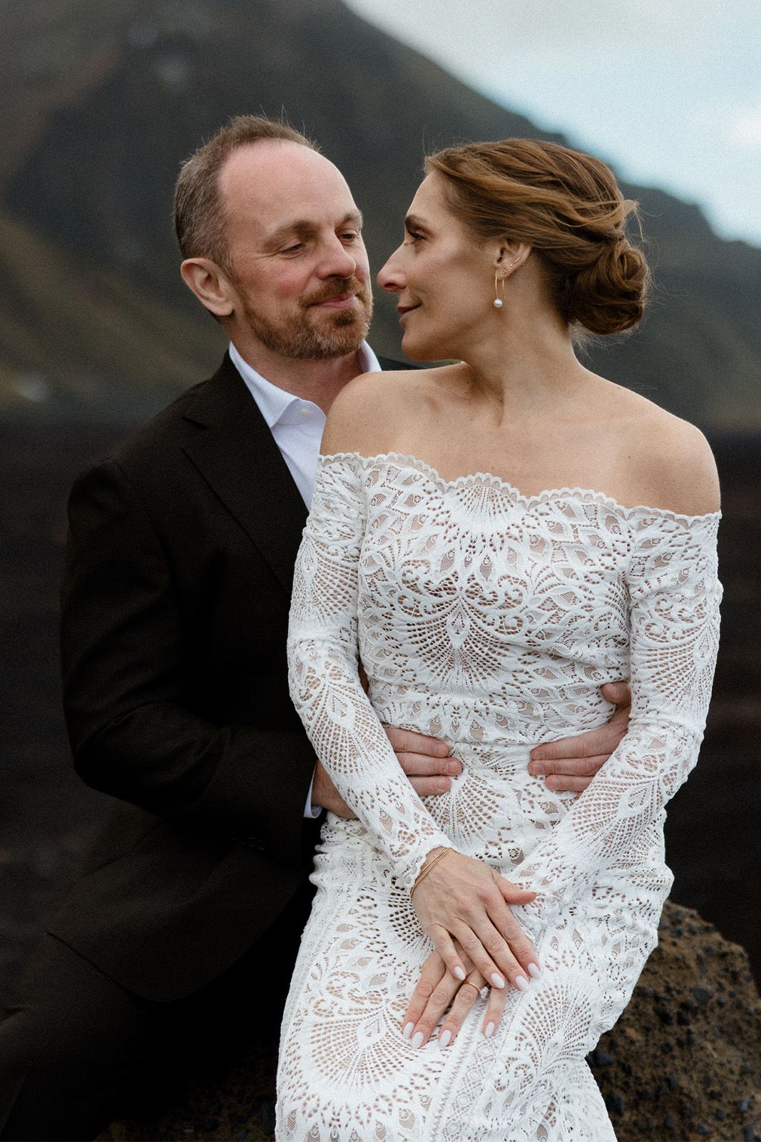 Couple embracing outdoors, wearing formal attire, with a scenic background.