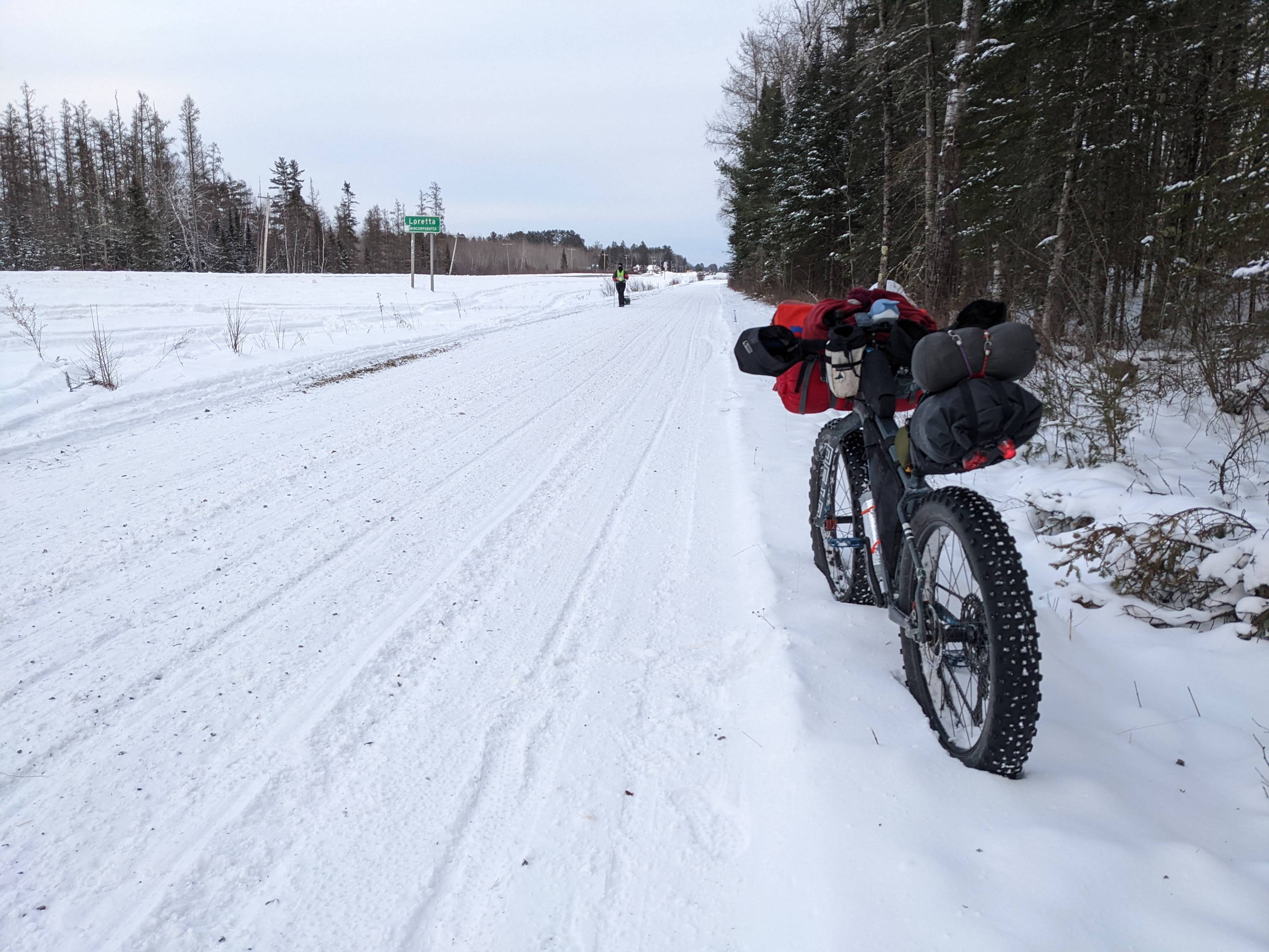 A fully-loaded bikepacking bike leans in the snow by a plowed trail road.