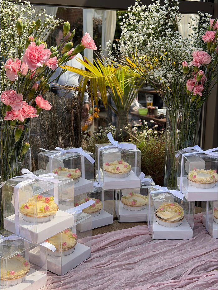 Cakes in clear boxes surrounded by pink flowers and greenery.