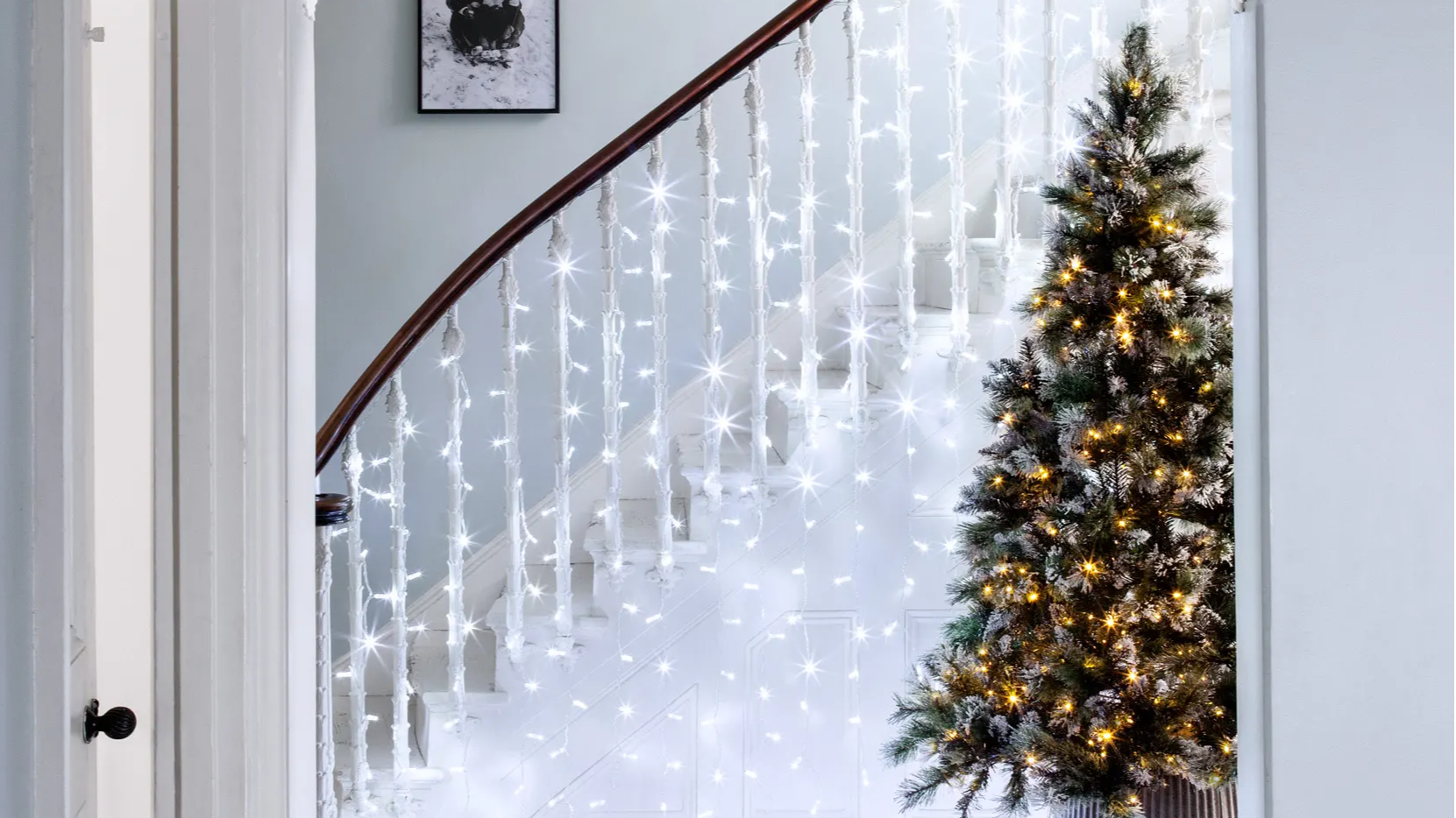 White curtain light displayed on staircase and hung from bannister with Christmas tree lit up alongside the staircase in the hallway