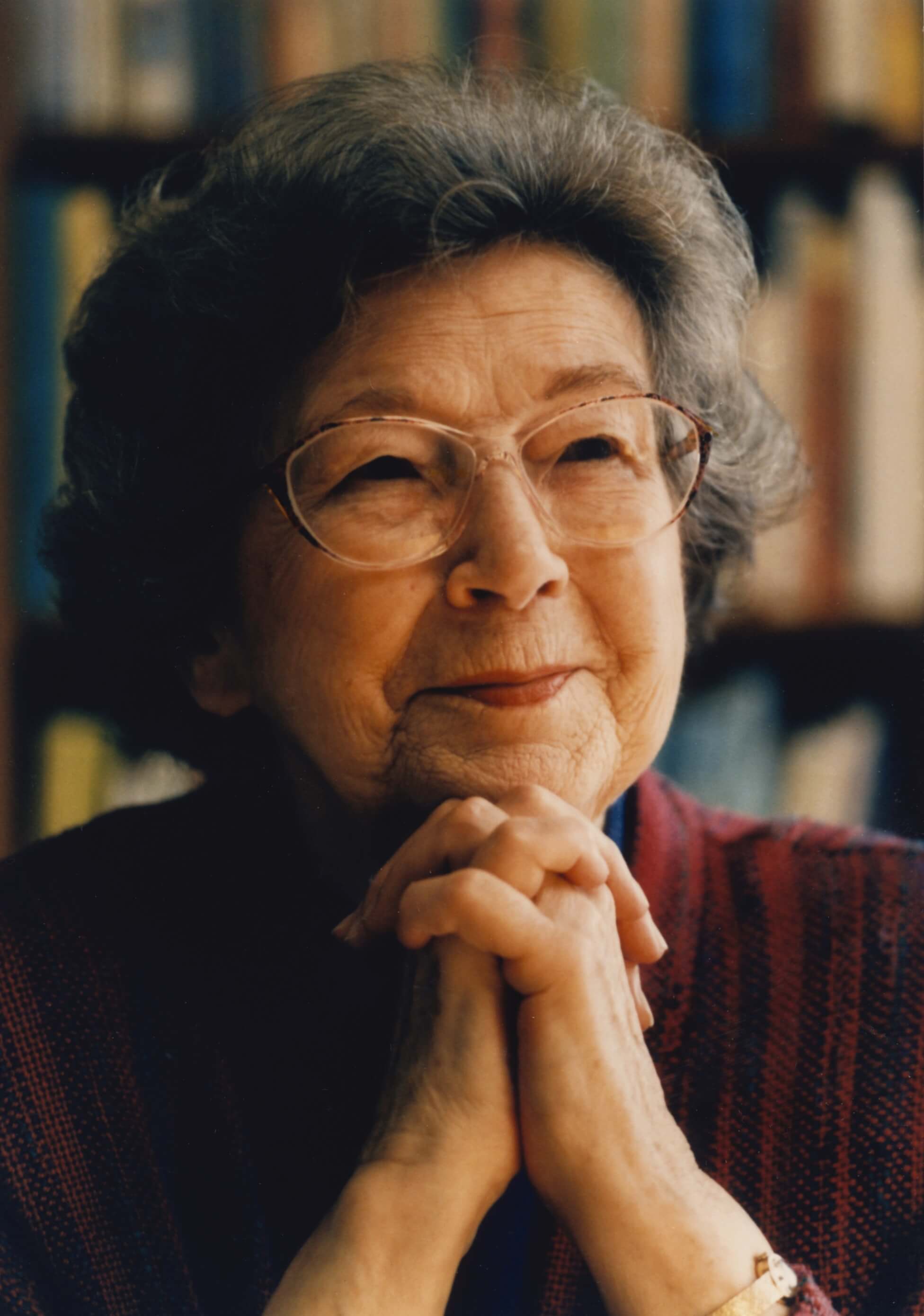 Elderly woman with glasses, smiling and resting chin on clasped hands.