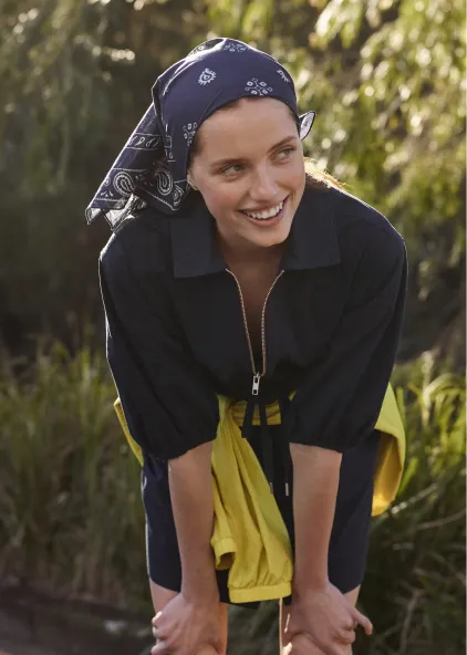 Smiling woman in a black outfit and blue bandana by greenery.