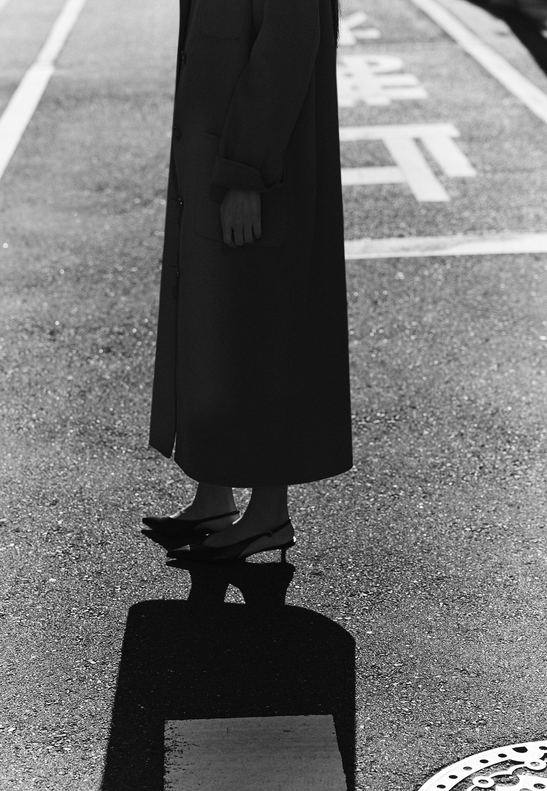 A person in a long coat stands on wet pavement, casting a shadow.