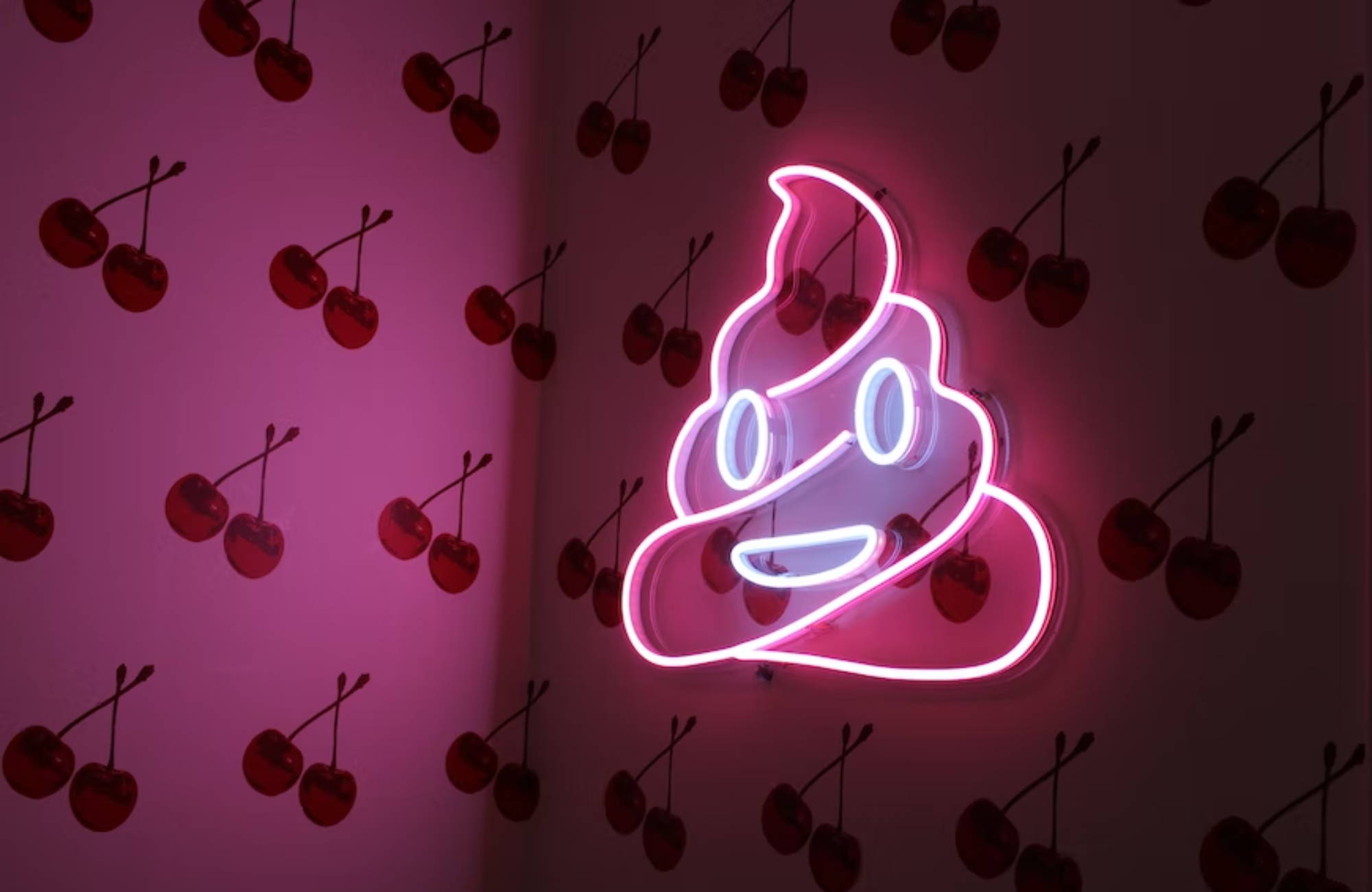 A neon pink poop emoji sign hanging on a wall with pink, cherry-print wallpaper.
