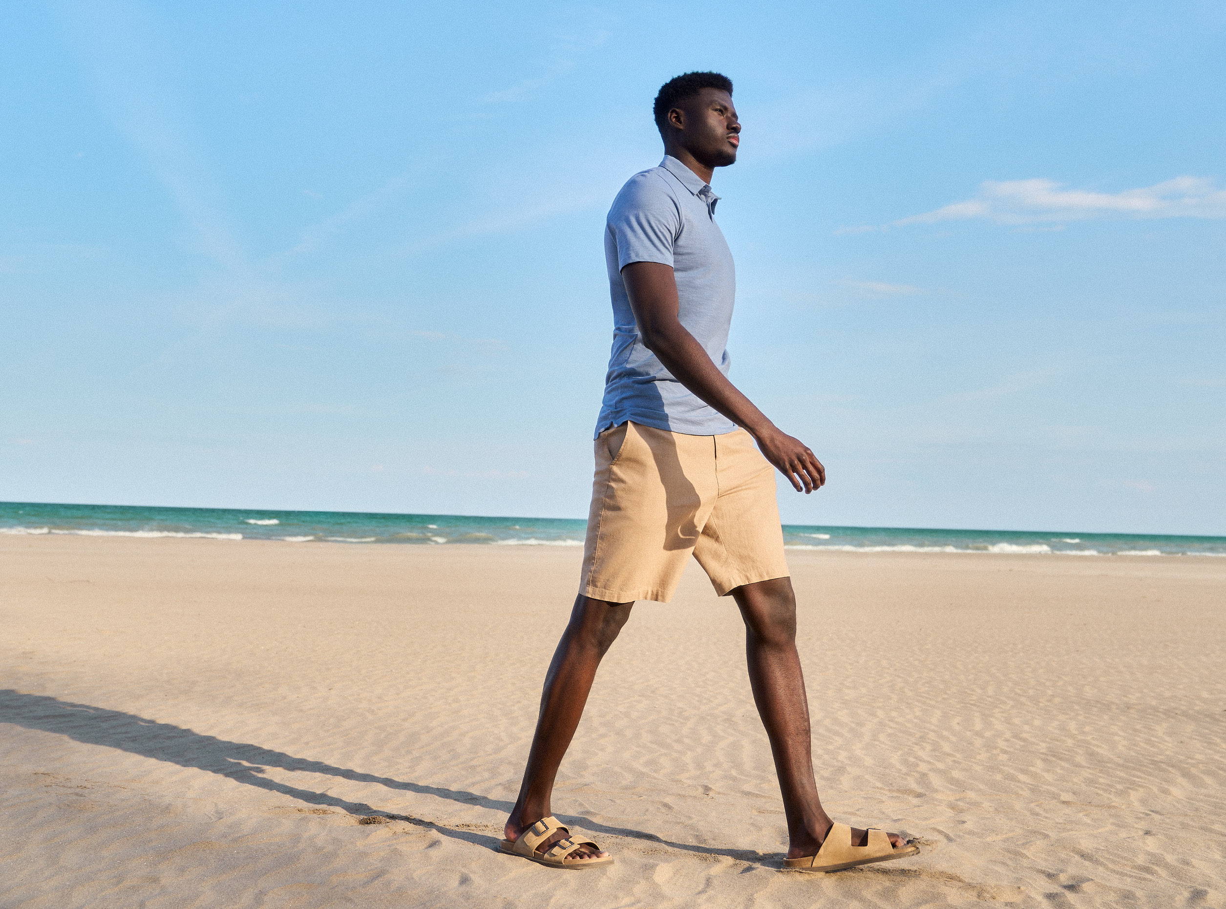 Tall man walking on the beach in a blue polo shirt and beige shorts