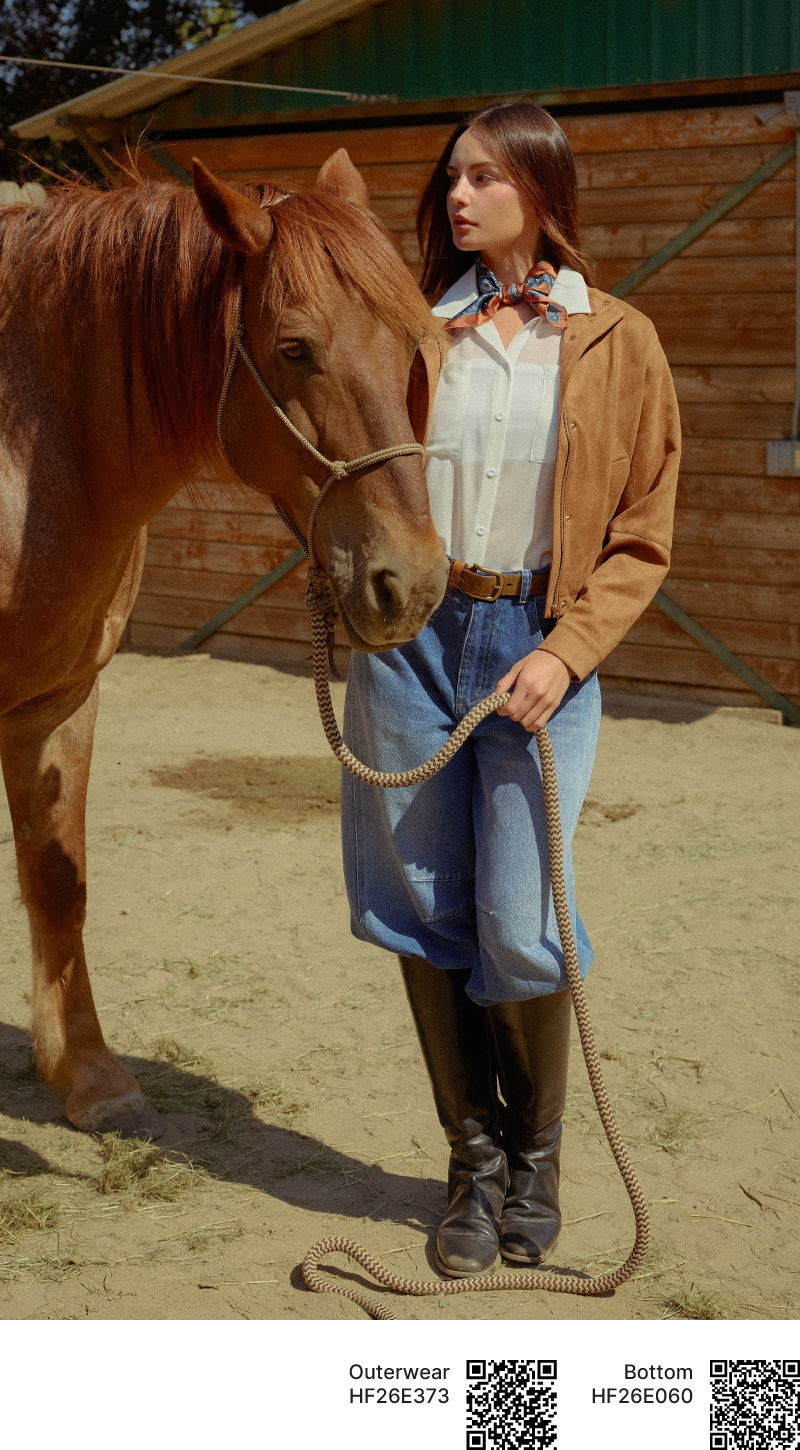 A woman in western attire stands beside a horse in a stable.