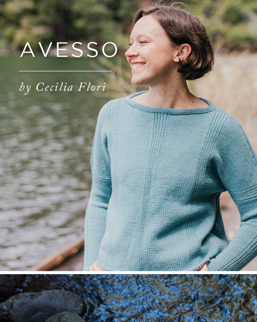 A woman stands on a lakeshore with her hand on her hip. She models a hand knit sweater with the title AVESSO by Cecelia Flori.