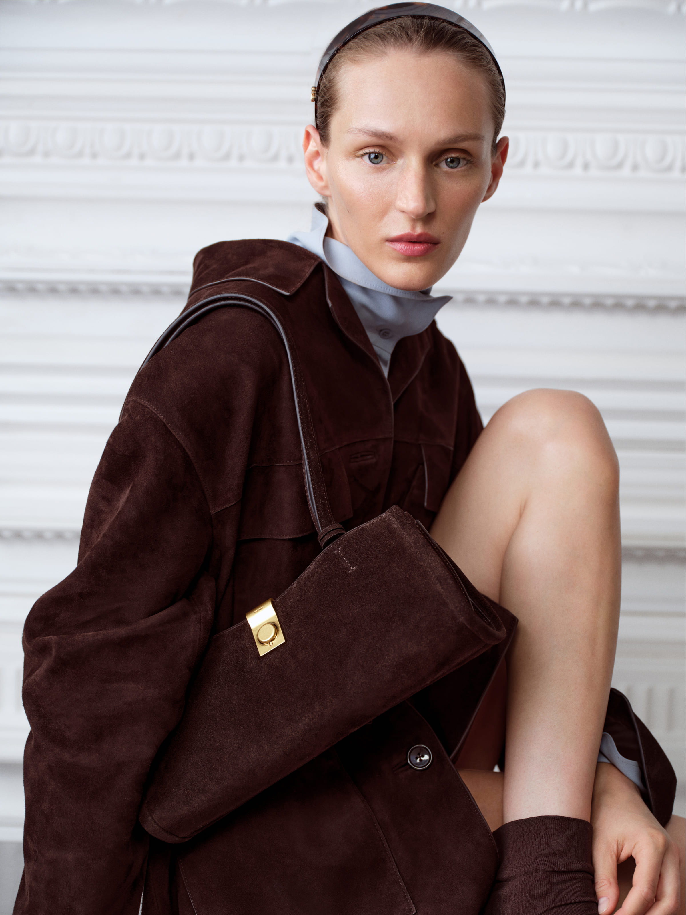 Model in a brown coat holding a stylish handbag, seated against a decorative background.