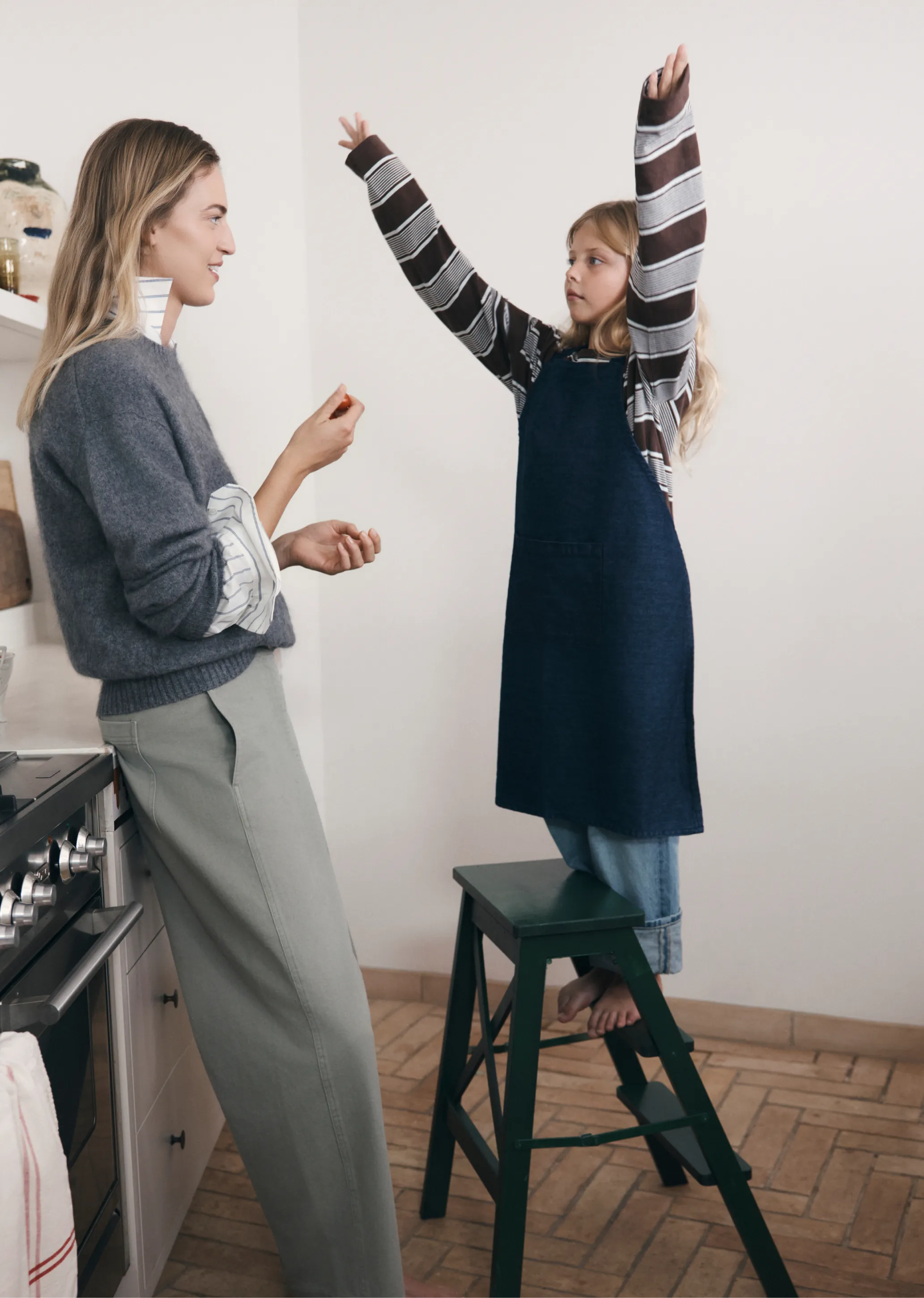 A woman and a girl interact in a kitchen, with the girl on a stool.