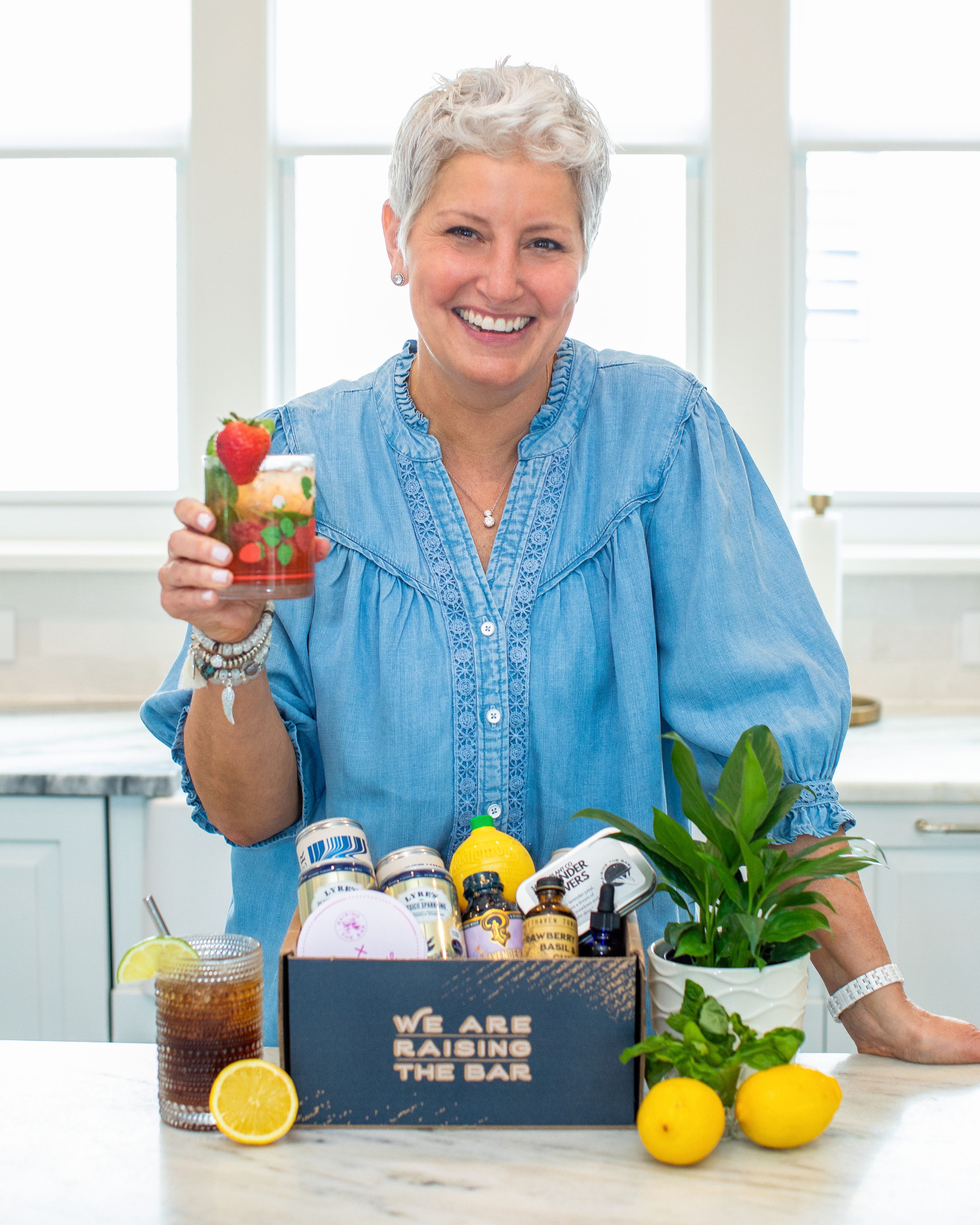 Mocktail Mom holding a strawberry mocktail in a bright kitchen in front of a box filled with drink making ingredients. 