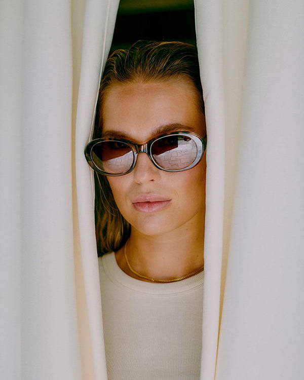 A woman looking out from behind cream curtains wearing the Amna sunglasses in Demi Tortoise.