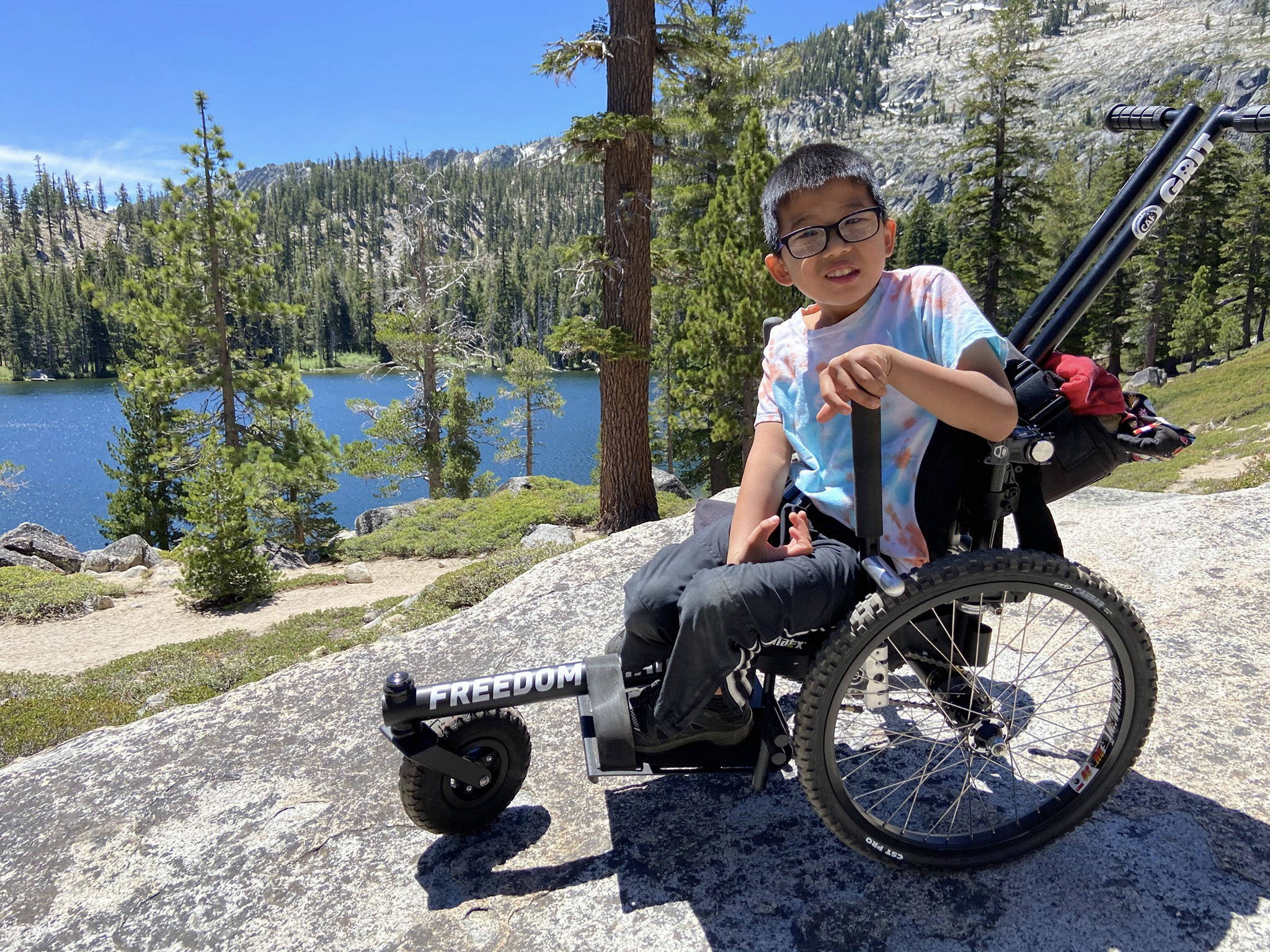 Child using GRIT Freedom Chair outdoor wheelchair on rocky slope with forest and water background