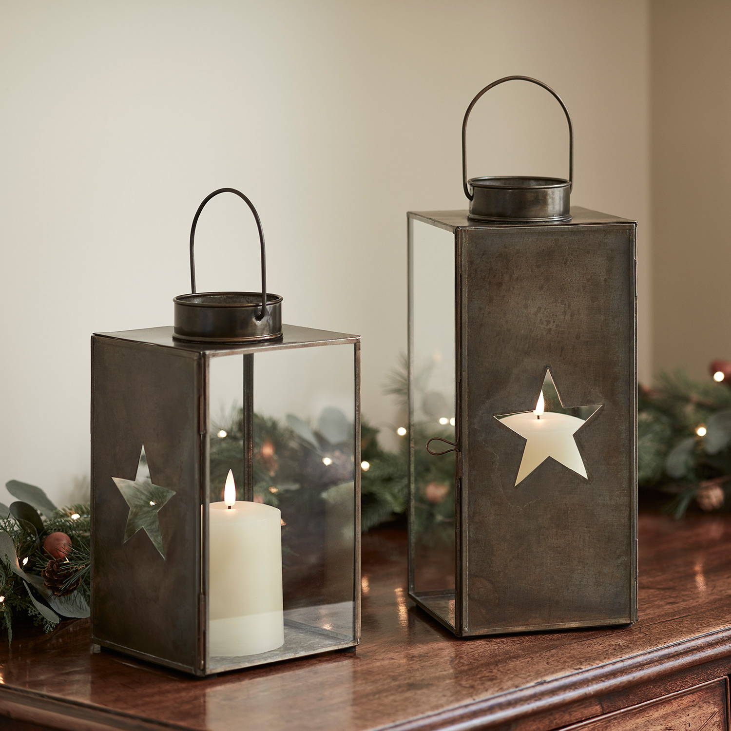 Brass star glass lantern & TruGlow® candles on a wooden cabinet with a Christmas garland.