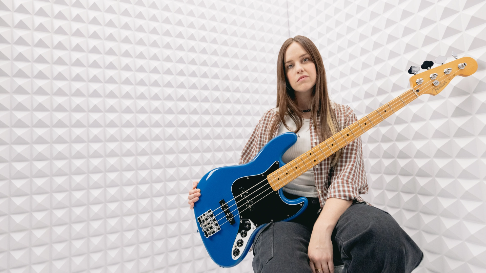 A woman sitting with a blue Fender Player II Modified Series Jazz Bass guitar in a soundproof room.
