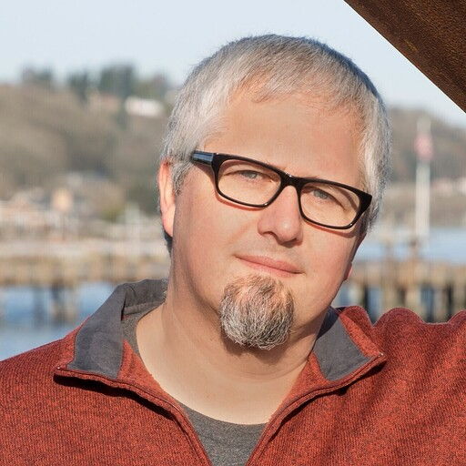 Man with glasses and a beard, wearing a red sweater by the water.