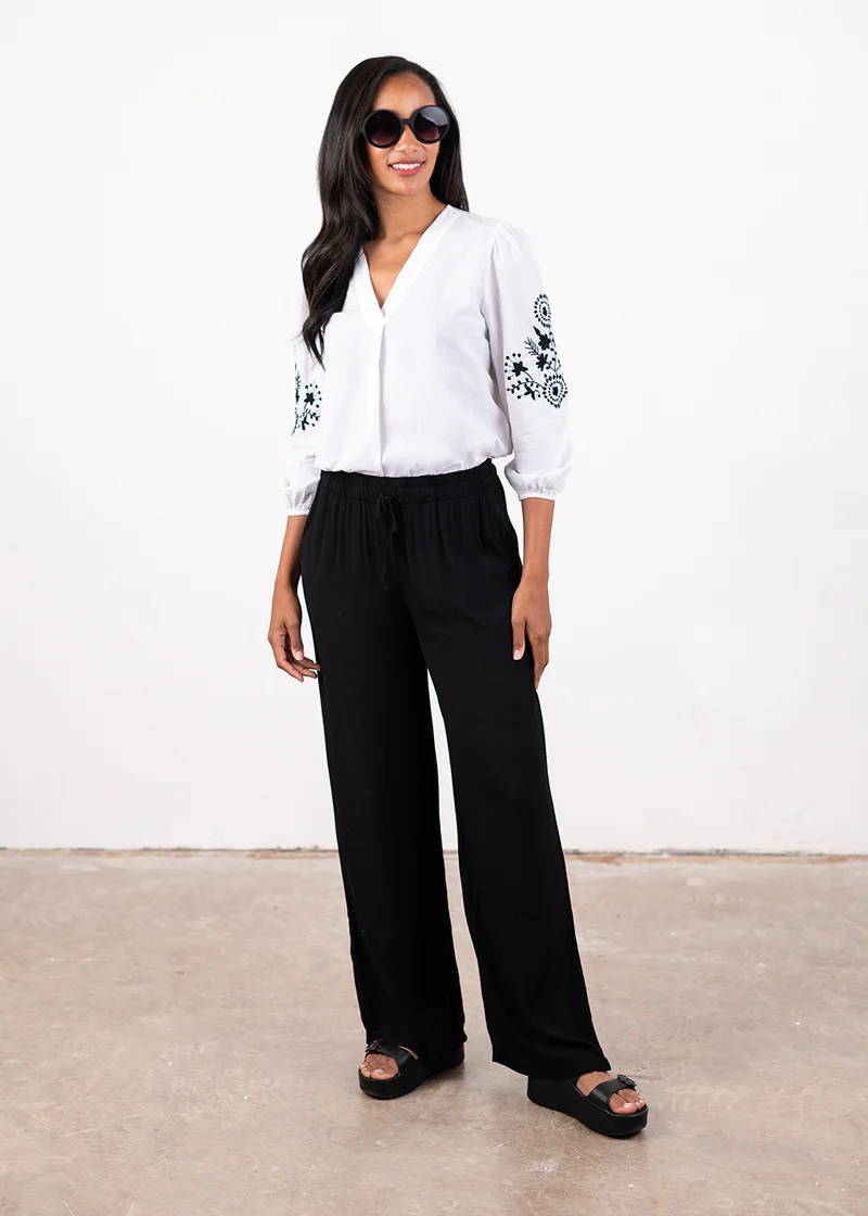 A model wearing a white shirt with black embroidered detailing on the sleeves, black wide leg trousers and black chunky platform slides