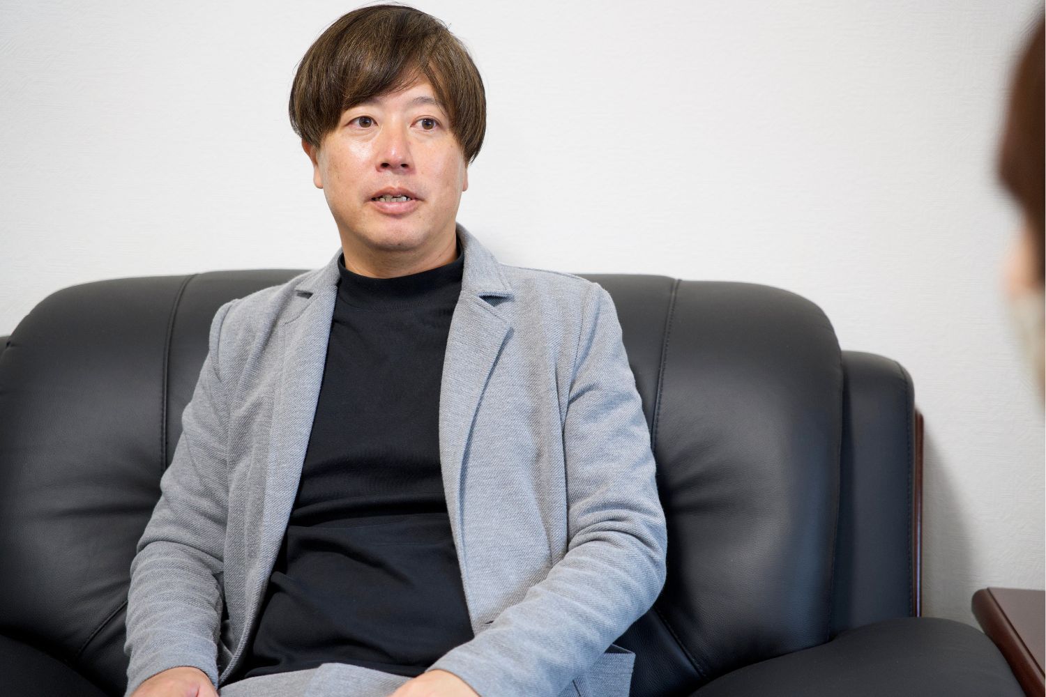 A person sitting on a couch, wearing a gray suit and black shirt.