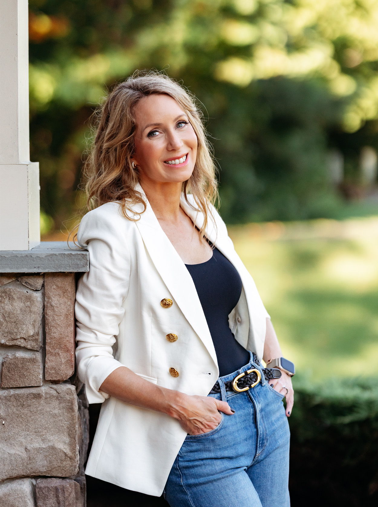 Woman smiling in a white blazer and jeans, leaning against a stone wall.