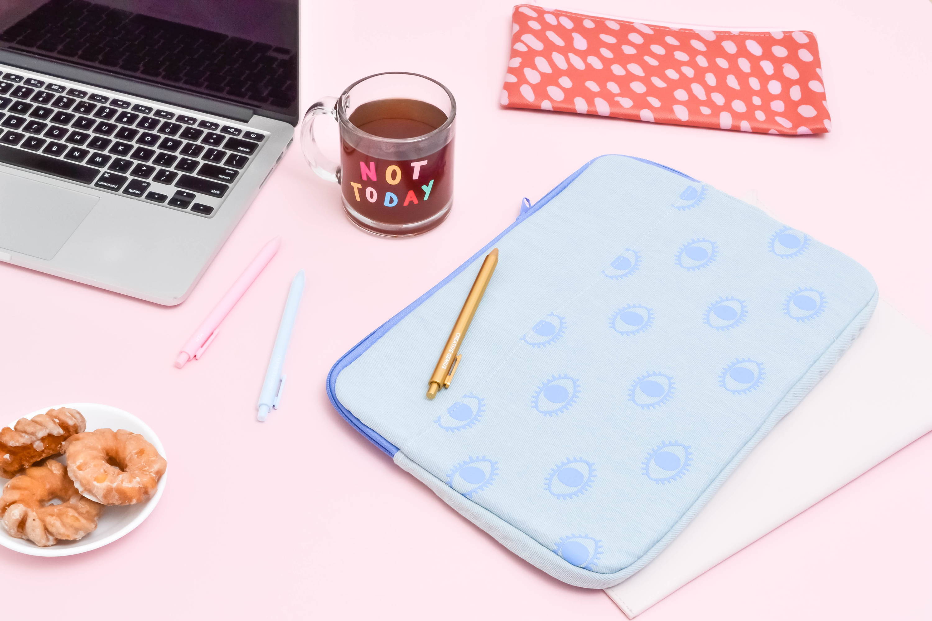 cute lifestyle image of a laptop, donuts, blue canvas laptop sleeve with eyeballs, a red vegan leather pouch, and a clear glass mug that says not today in rainbow letters 