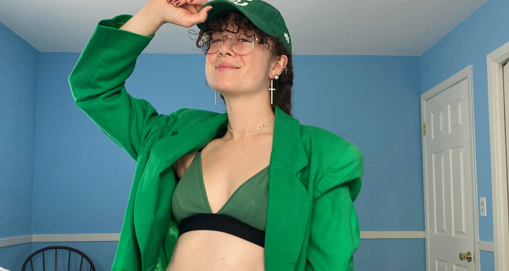 woman in a green blazer, green bralette, and green baseball hat smiles and holds her baseball hat