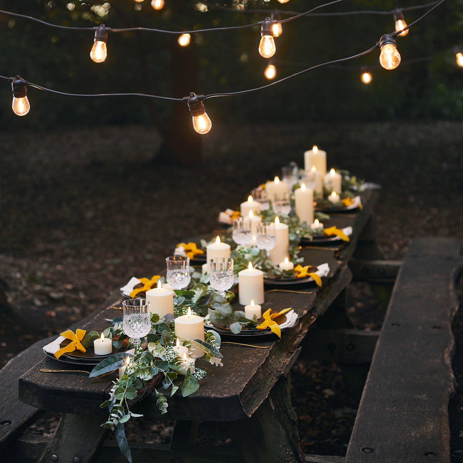 Warm white festoon lights over an outdoor wedding dining table with LED candles and a green garland.