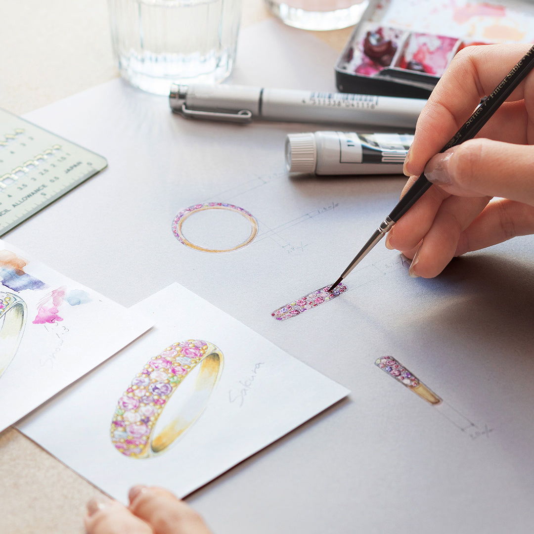 A hand painting jewelry designs on paper with tools and materials nearby.