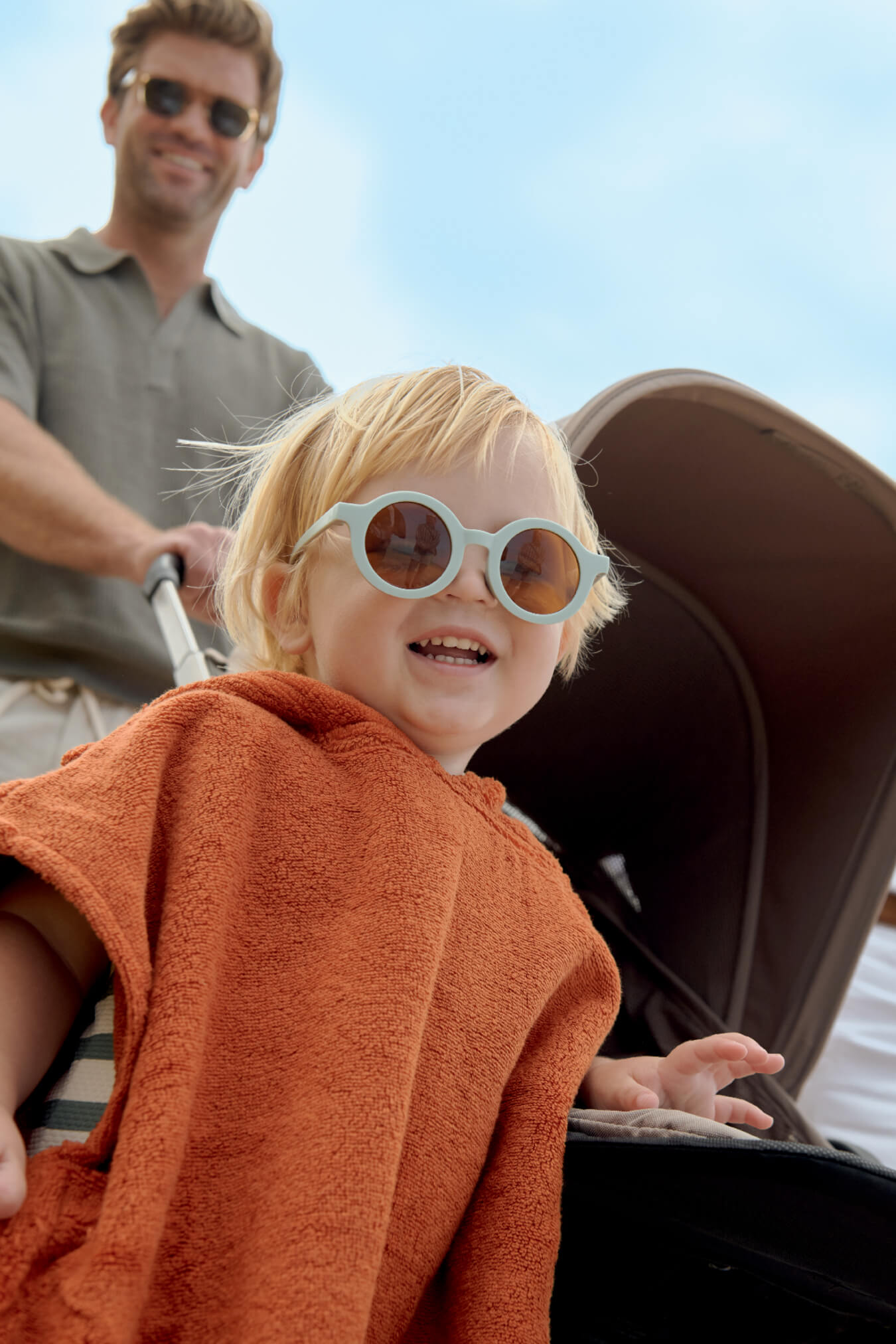 Child in sunglasses smiling with a stroller in the background.