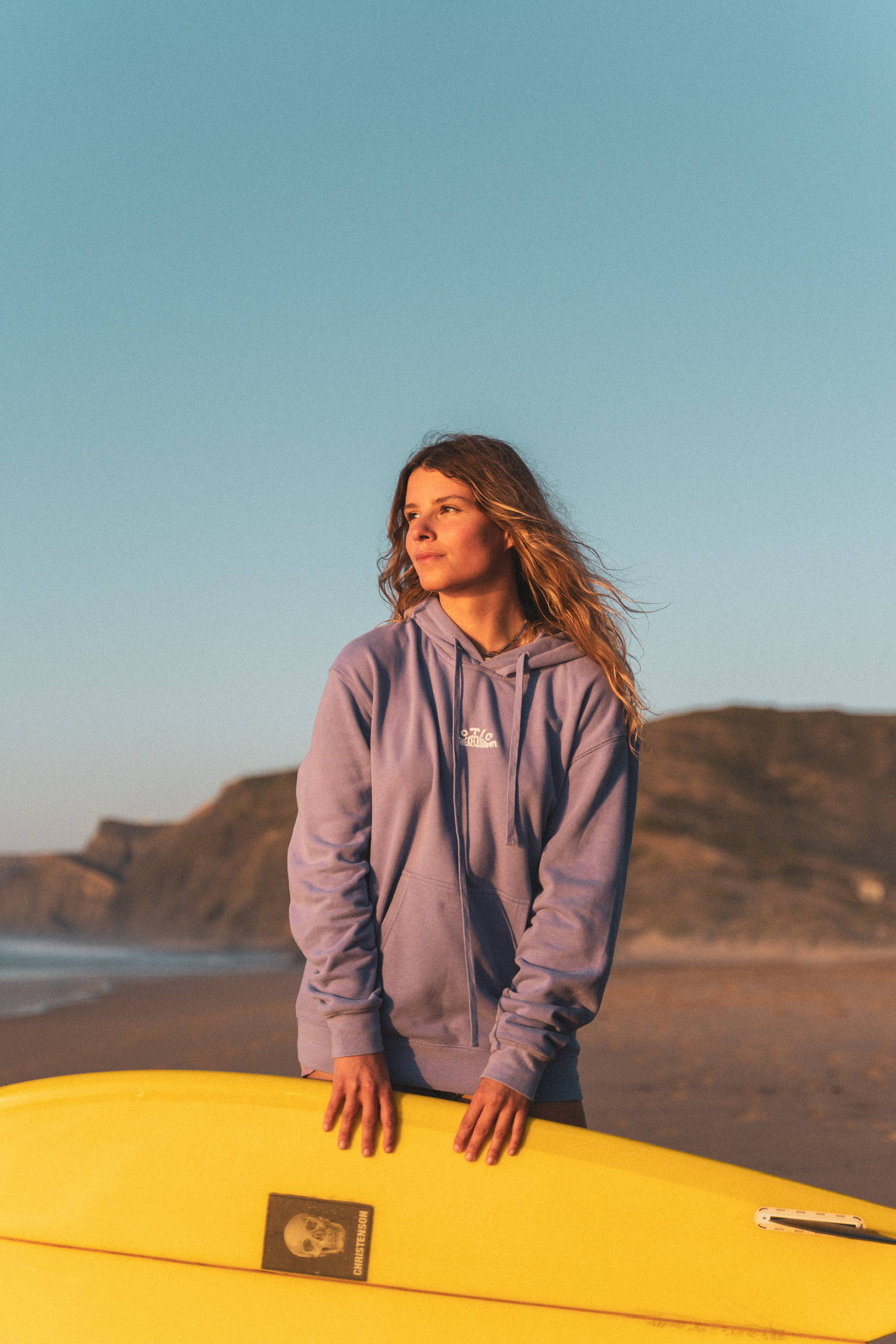 A woman wearing a lilac purple hoodie holding her yellow surfboard on a beach in France