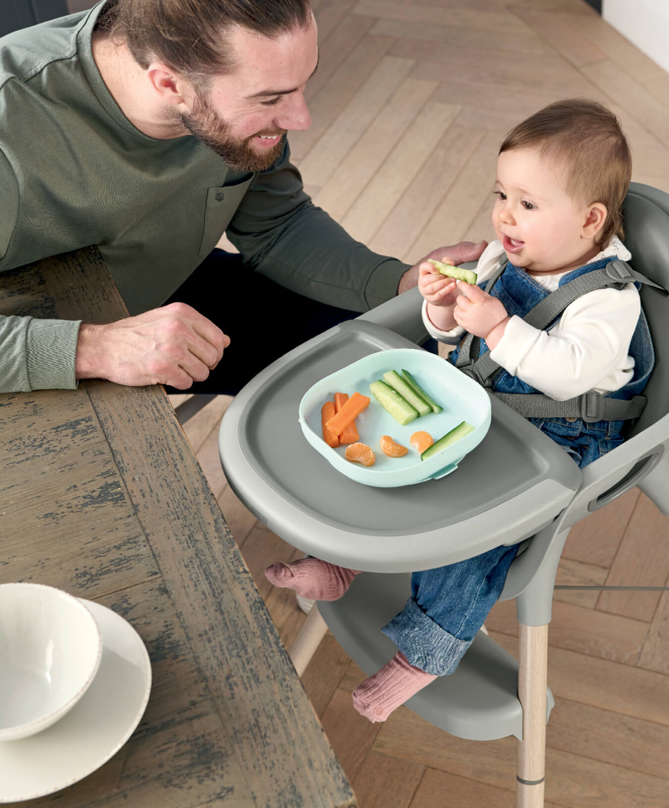 A father feeding his baby in a high chair at a table.
