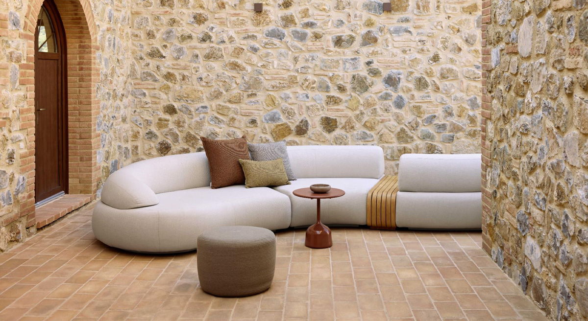 Curved white Cane-line sofa with cushions in a stone-walled room.