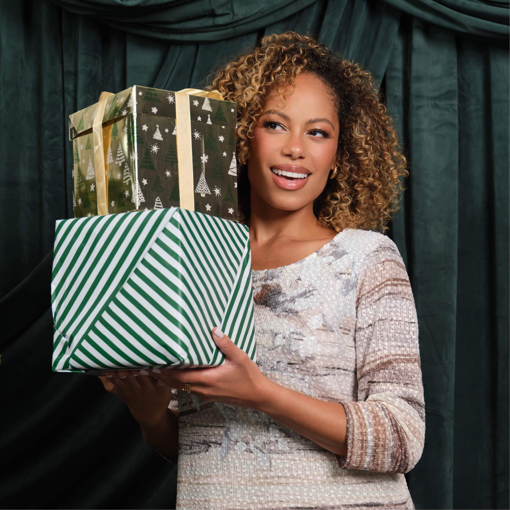 Smiling woman holding wrapped gifts in front of a dark green backdrop.