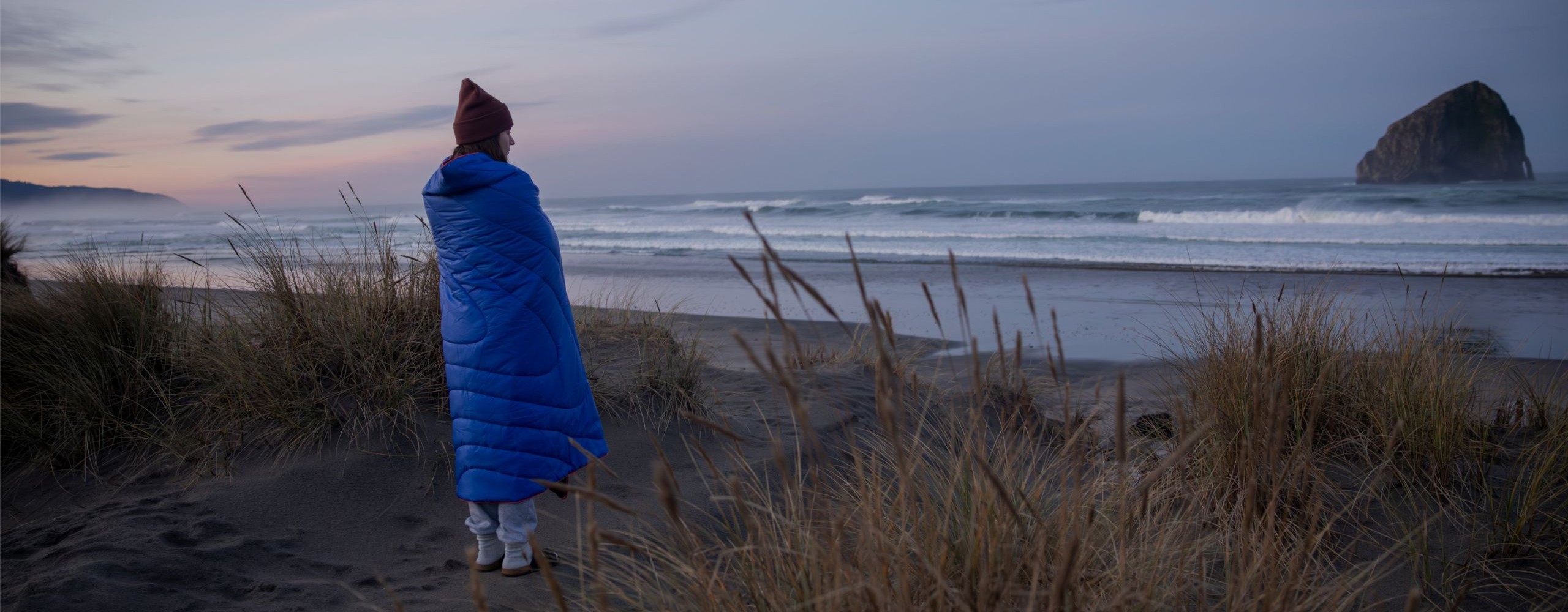 Person in a blue blanket standing on a beach at dusk.
