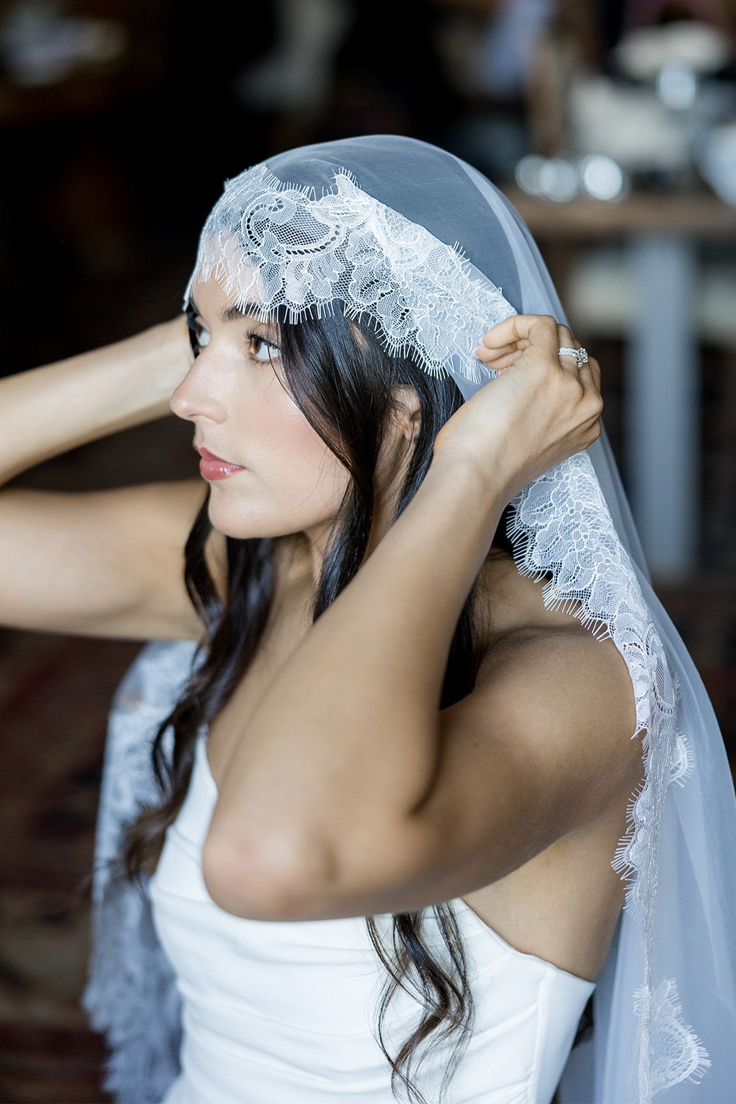 Bride adjusting her lace veil in an indoor setting.