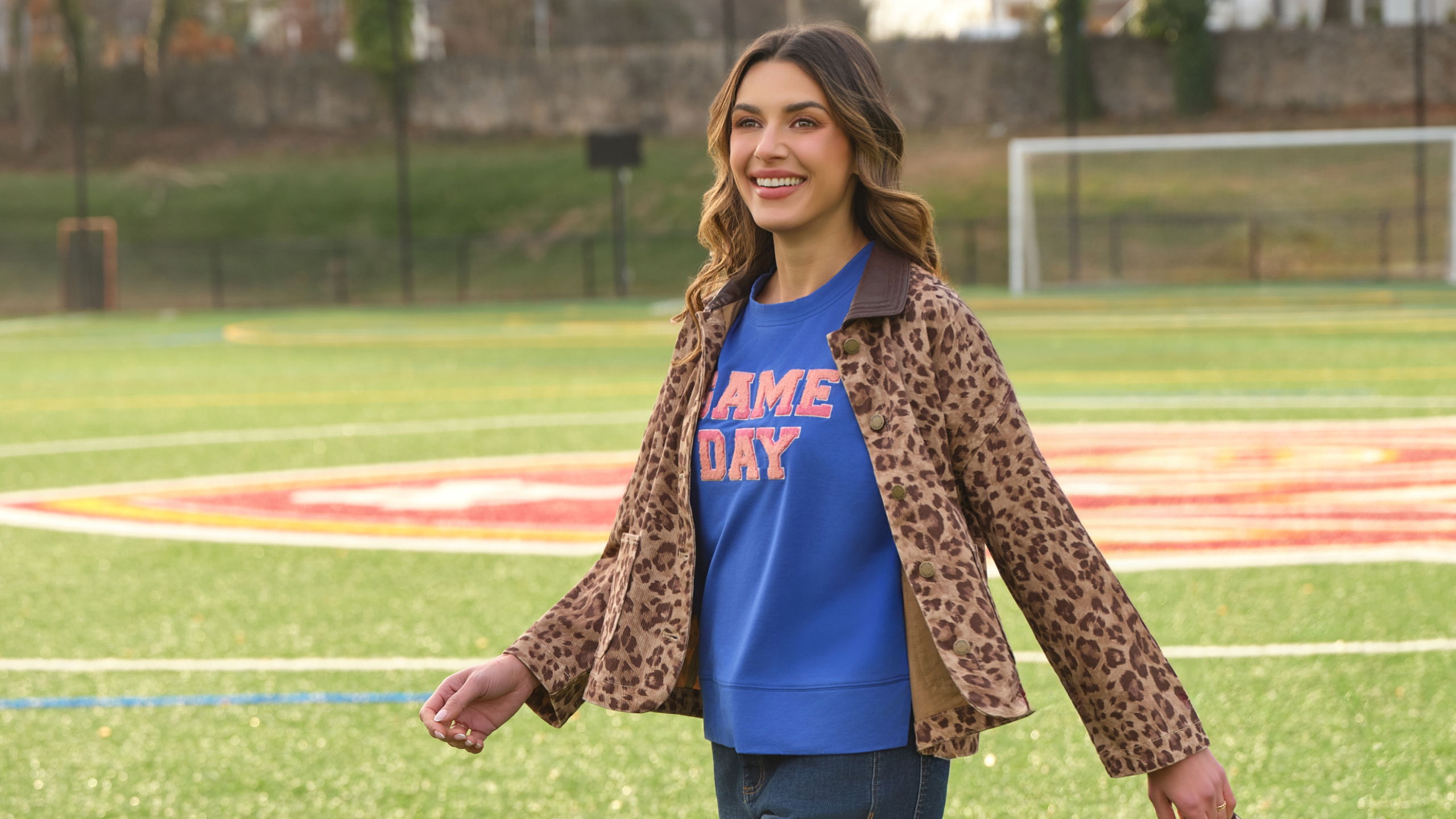 Person walking on a soccer field, wearing a blue shirt and leopard print jacket.