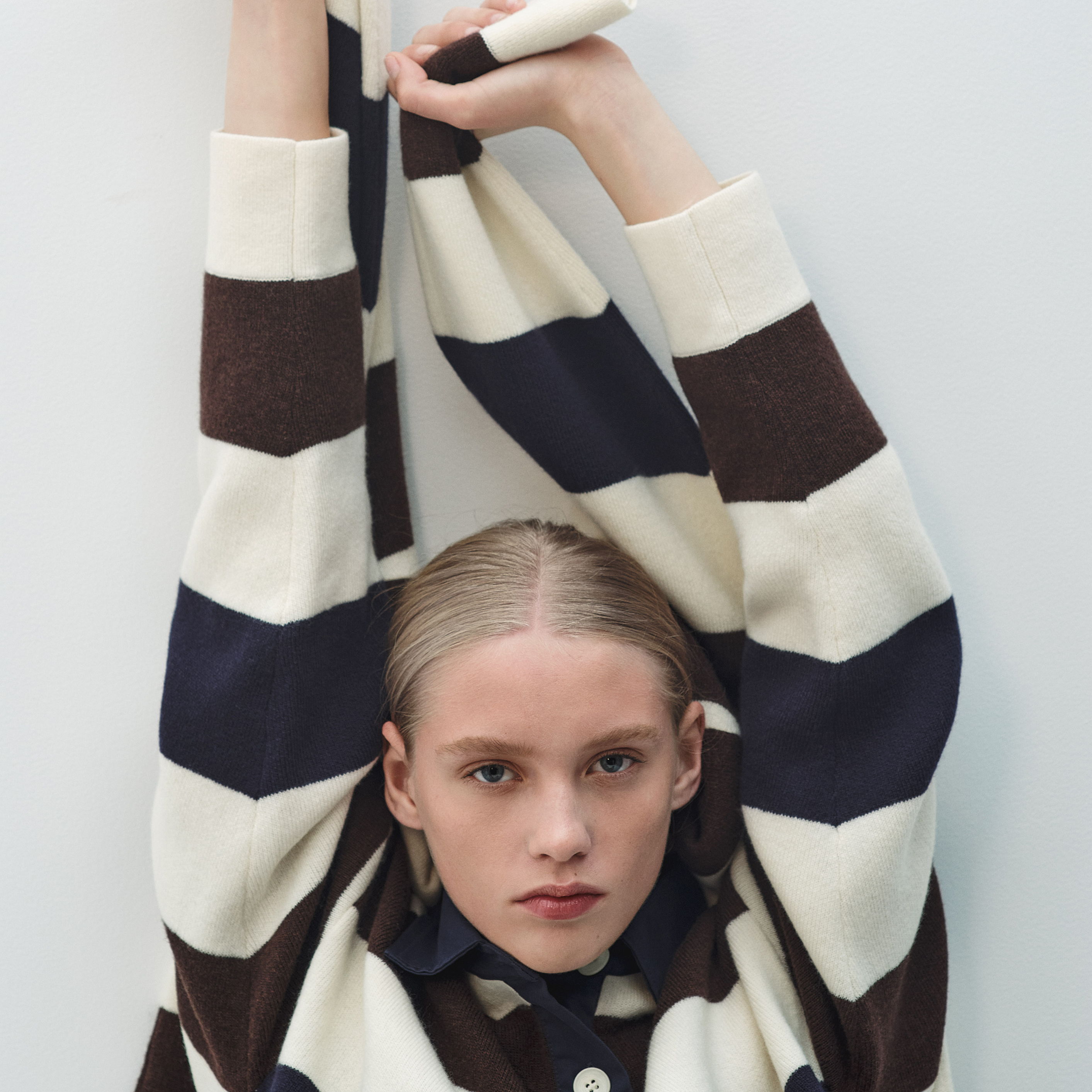 A person in a striped sweater poses with arms raised against a plain background.