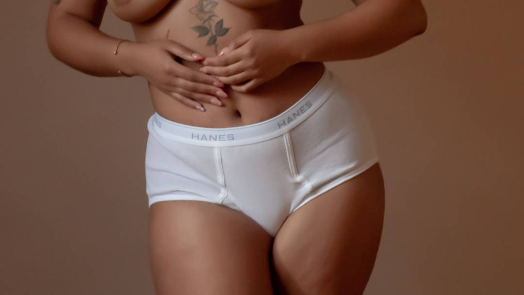 Image of woman's torso with tattoo from mid-thigh to under-bust wearing briefs and  with  hands placed over abdomen.